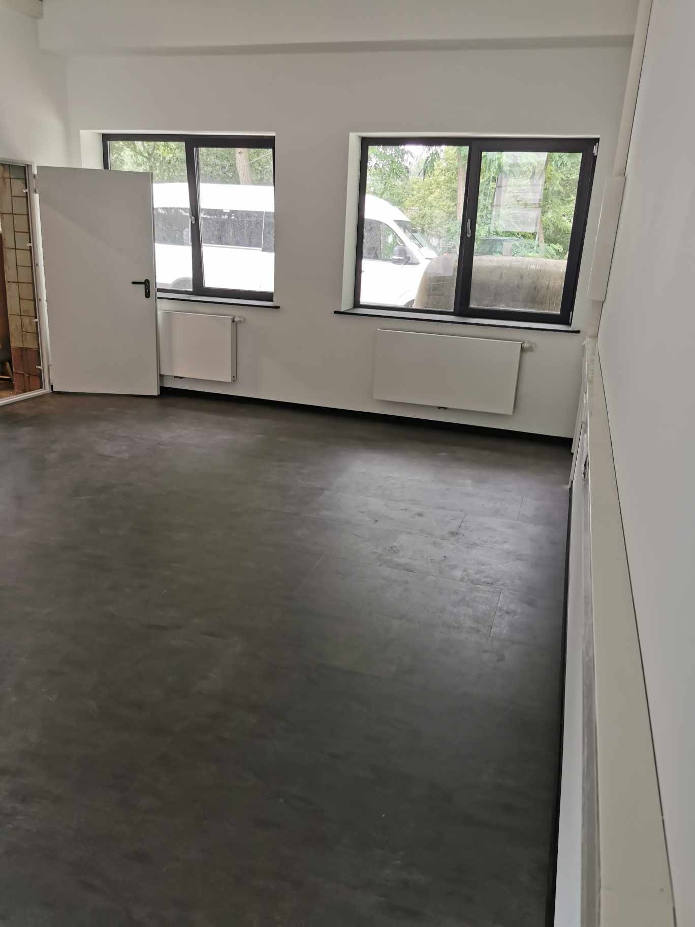 Wnętrze odnowionego biura z szarą podłogą, białymi ścianami, grzejnikami pod oknami i otwartymi białymi drzwiami.