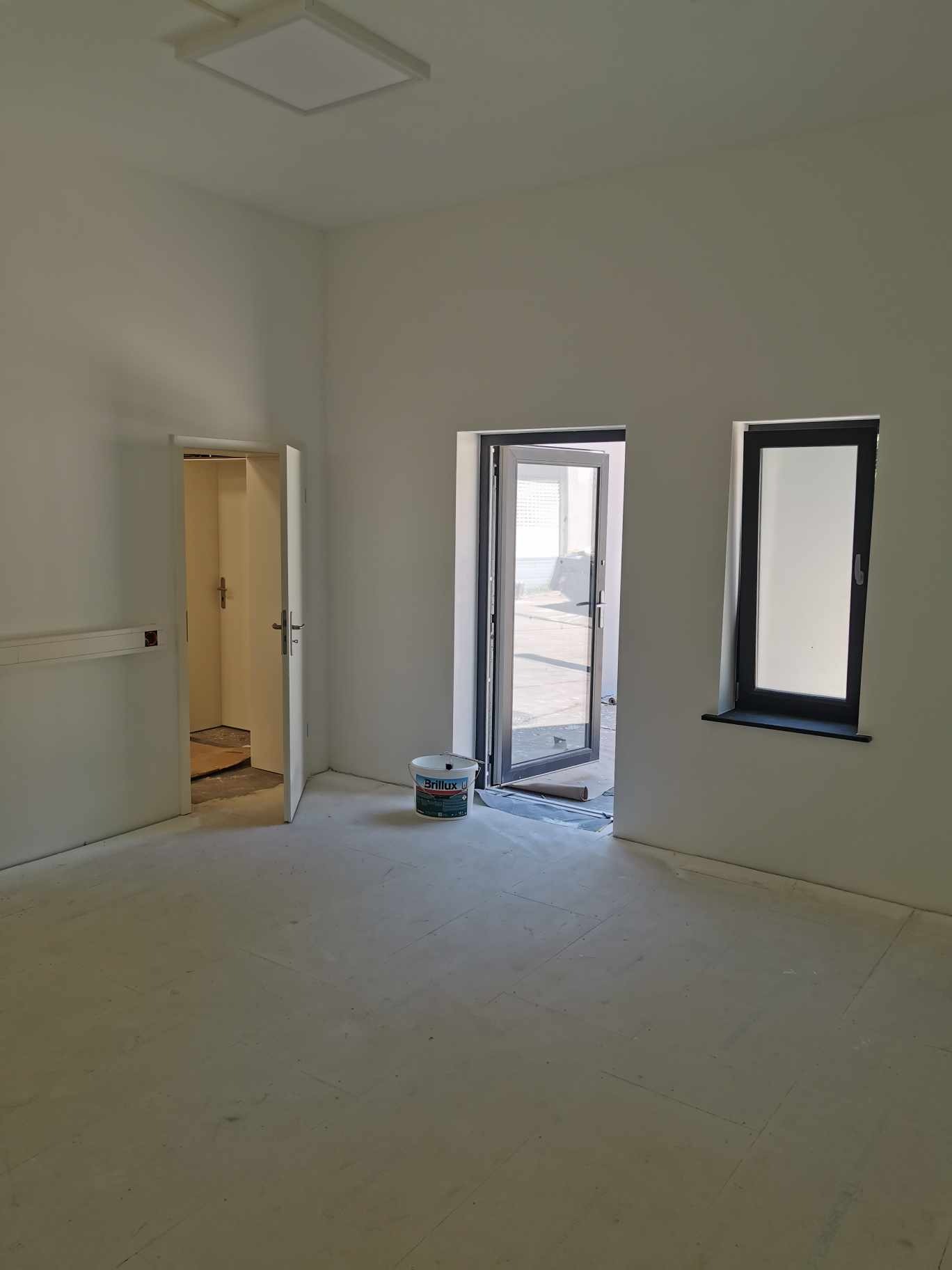 Wnętrze pokoju po remoncie, świeżo pomalowane ściany na biało, widoczne drzwi i okno z czarnymi ramami, na podłodze ślady farby, wiaderko farby marki Brillux.
