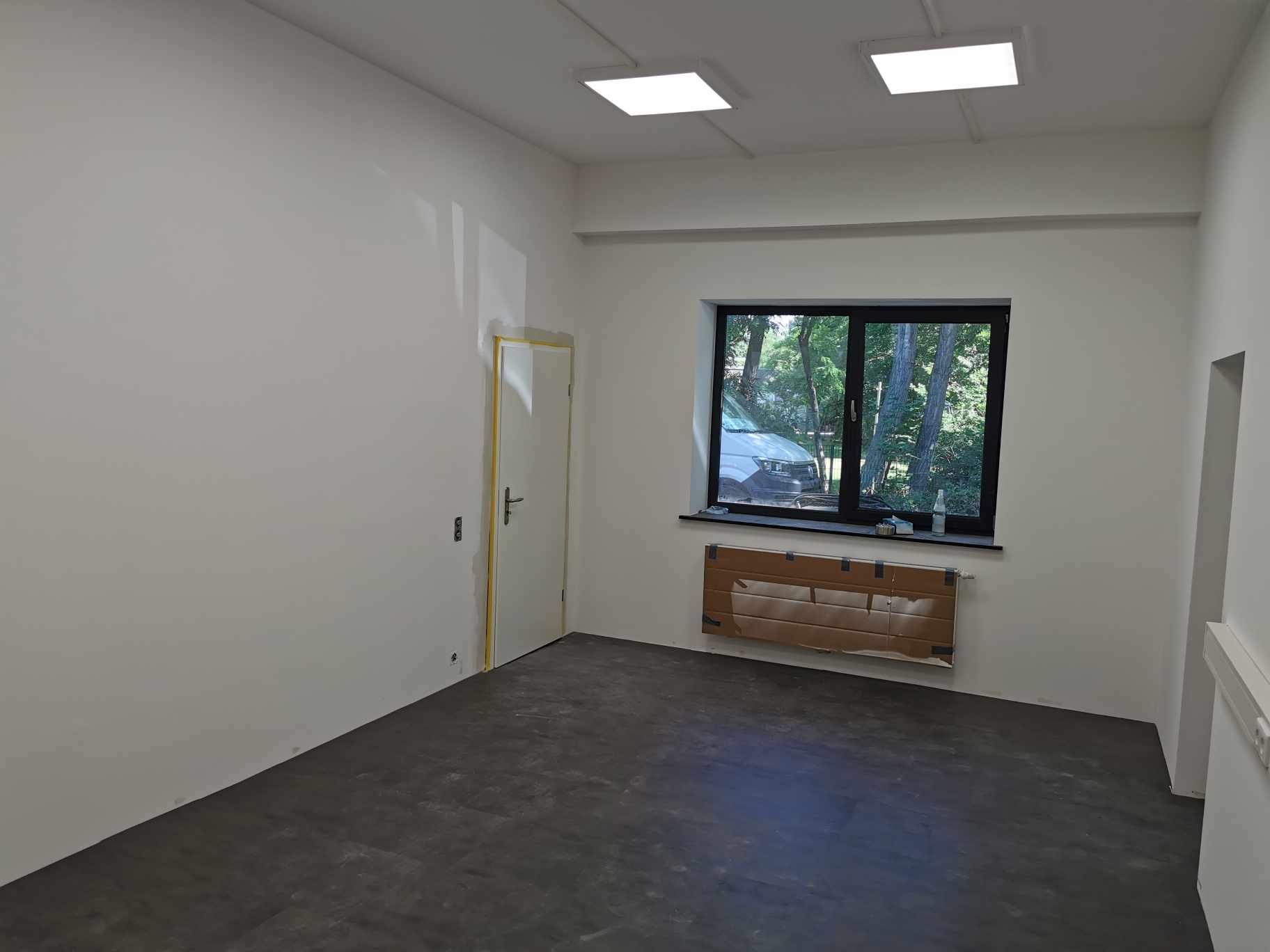 Wnętrze biura po świeżym pomalowaniu ścian na biało, z zabezpieczonym grzejnikiem kartonem i widokiem na zieleń przez okno.