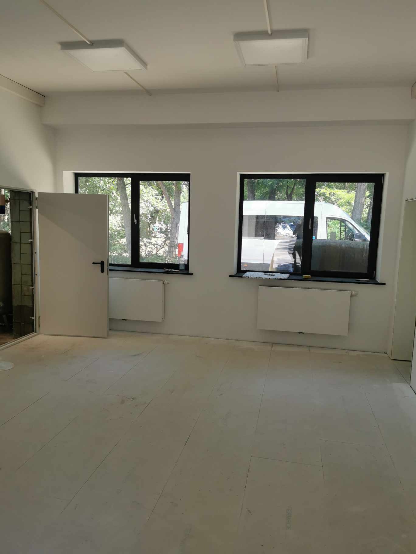 Wnętrze pustego, świeżo pomalowanego biura z dwoma oknami, białymi ścianami i grzejnikami, otwartymi drzwiami po lewej stronie i widocznym na zewnątrz samochodem dostawczym.