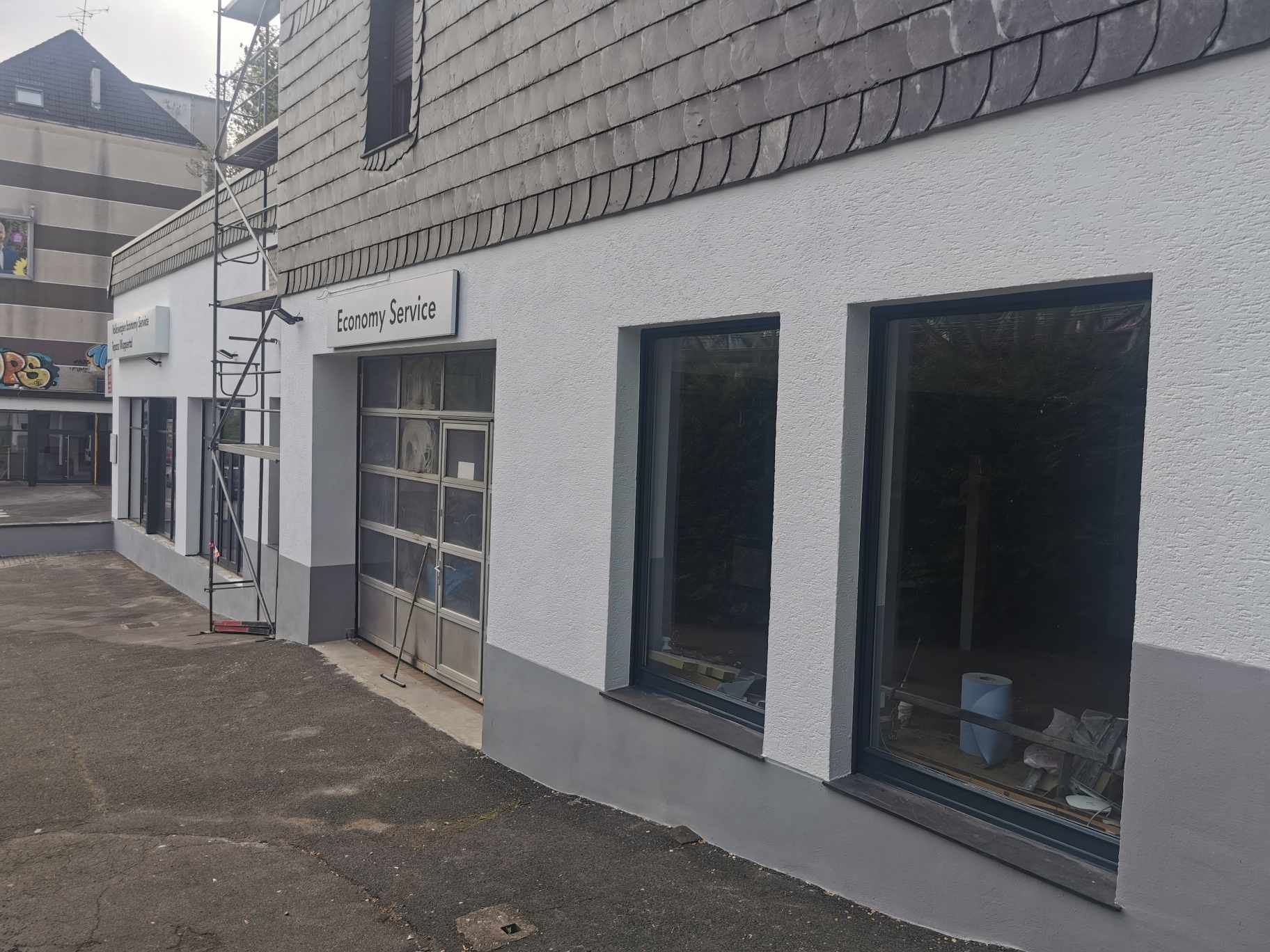Odnowiona elewacja budynku usługowego z szarą dachówką, widoczne rusztowanie i logo 'Economy Service', dolna część ściany pomalowana na szaro.