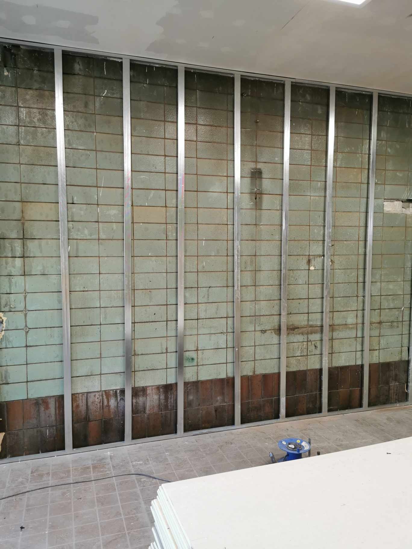 Montaż metalowej konstrukcji pod płyty gipsowo-kartonowe na starej, częściowo odrapanej ścianie z płytek w budynku we Wrocławiu. Na podłodze stos płyt gipsowych i narzędzia.