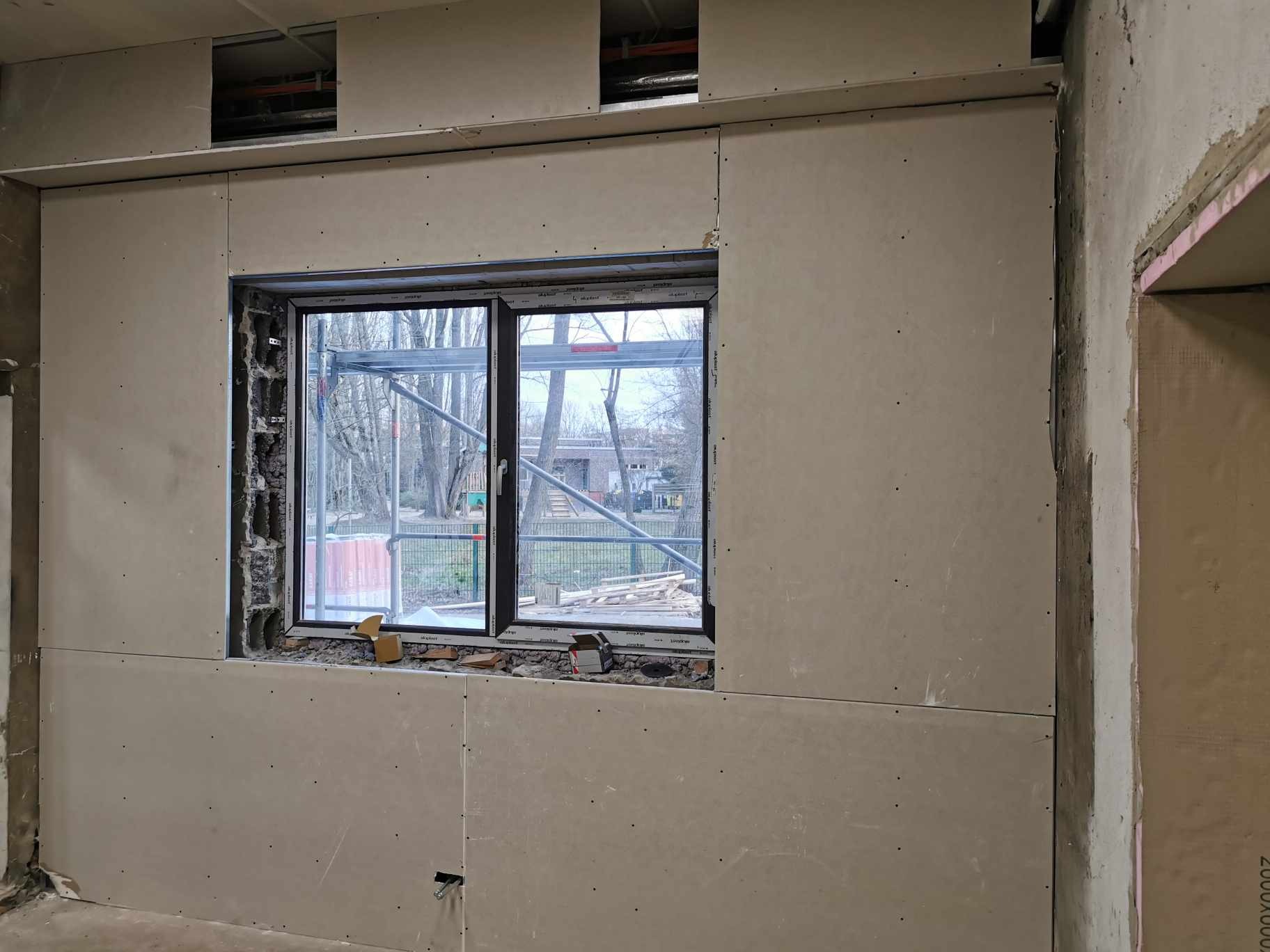 Montaż płyt kartonowo-gipsowych na ścianie wokół okna, widoczne szpachlowane łączenia i otwory montażowe, w tle rusztowanie za oknem.