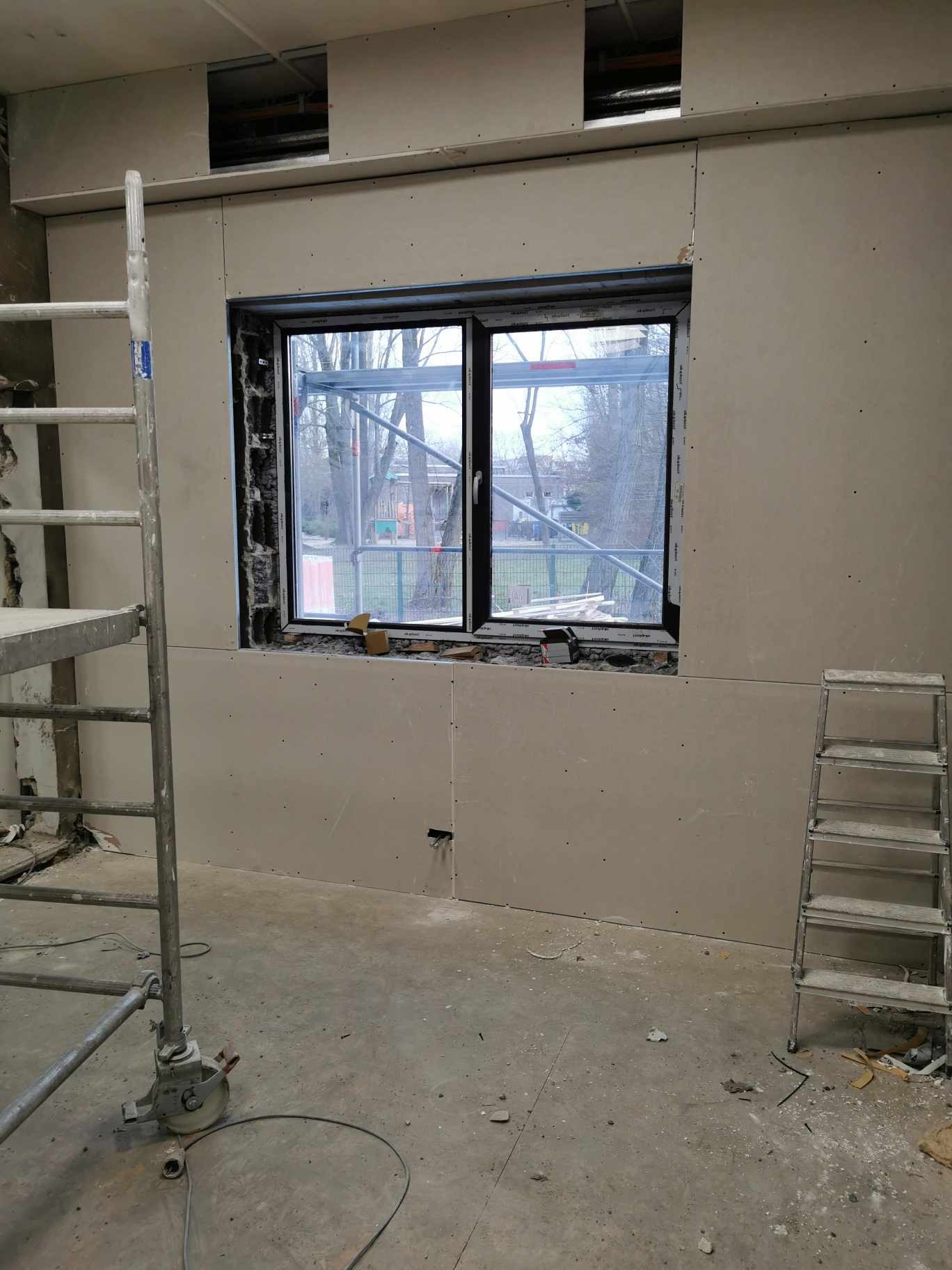 Montaż płyt kartonowo-gipsowych wokół okna, widoczny fragment rusztowania i drabina, surowy stan wykończenia wnętrza.