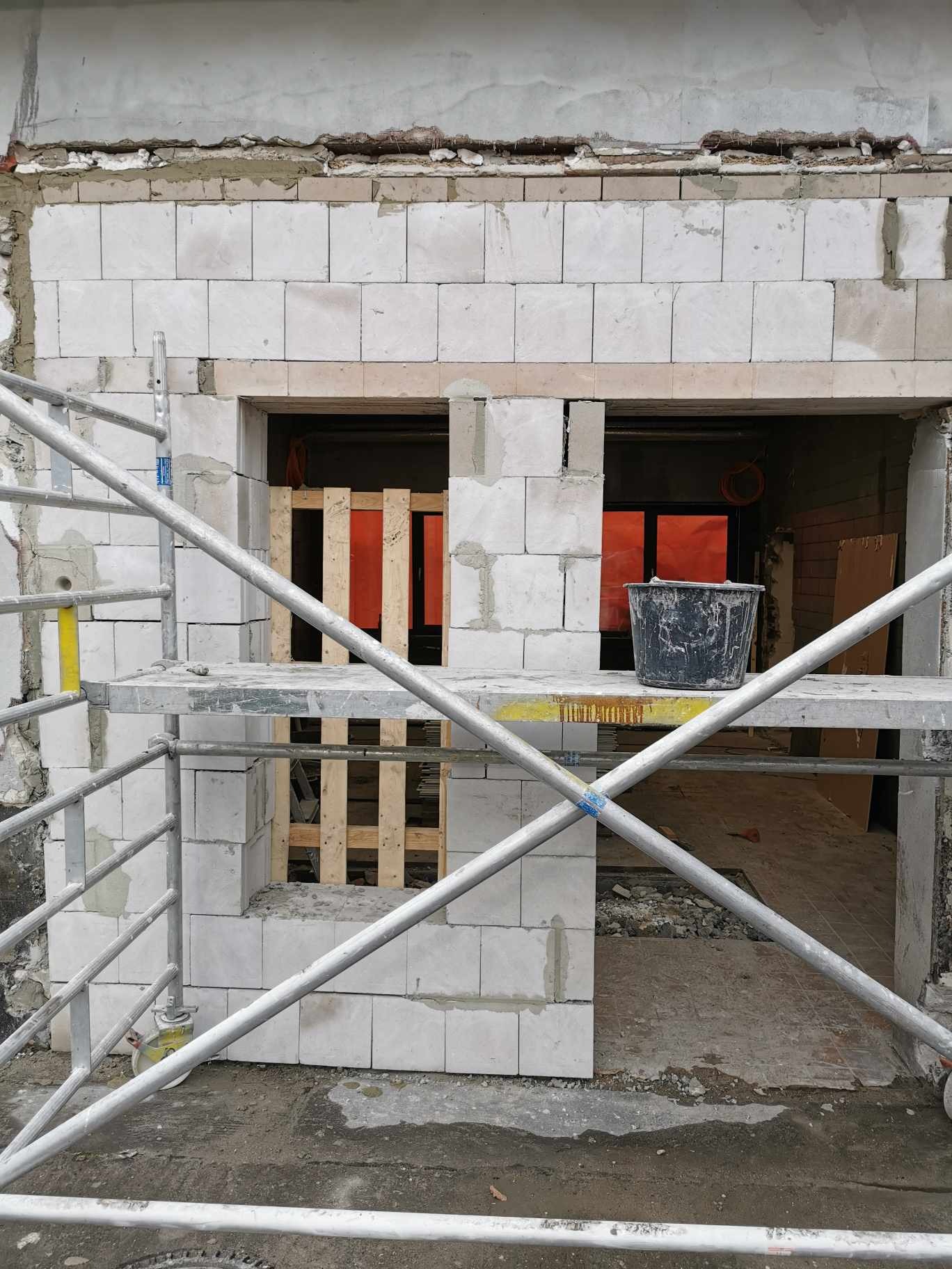Wznoszenie ściany z białych bloczków na zaprawie, widoczne rusztowanie, drewniane szalunki okienne i czarne wiadro na podeście.