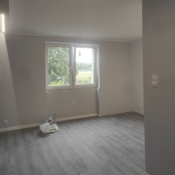 Pomalowane na szaro wnętrze pokoju z widokiem na zieleń za oknem. Na podłodze panele imitujące drewno. Widoczny worek z akcesoriami malarskimi.