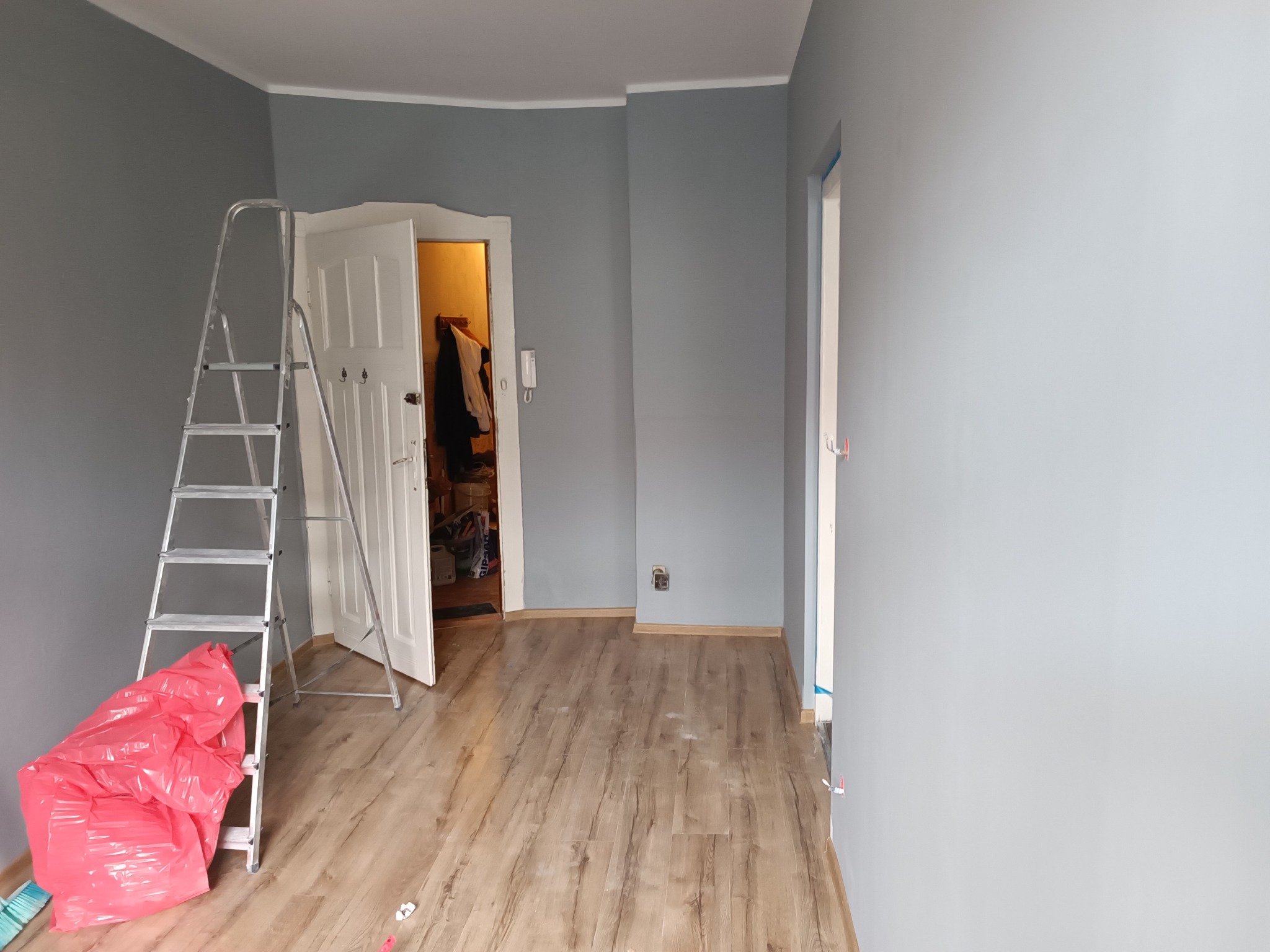 Świeżo pomalowane na szaro wnętrze pokoju z widoczną drabiną, workiem na śmieci i otwartymi drzwiami do innego pomieszczenia.