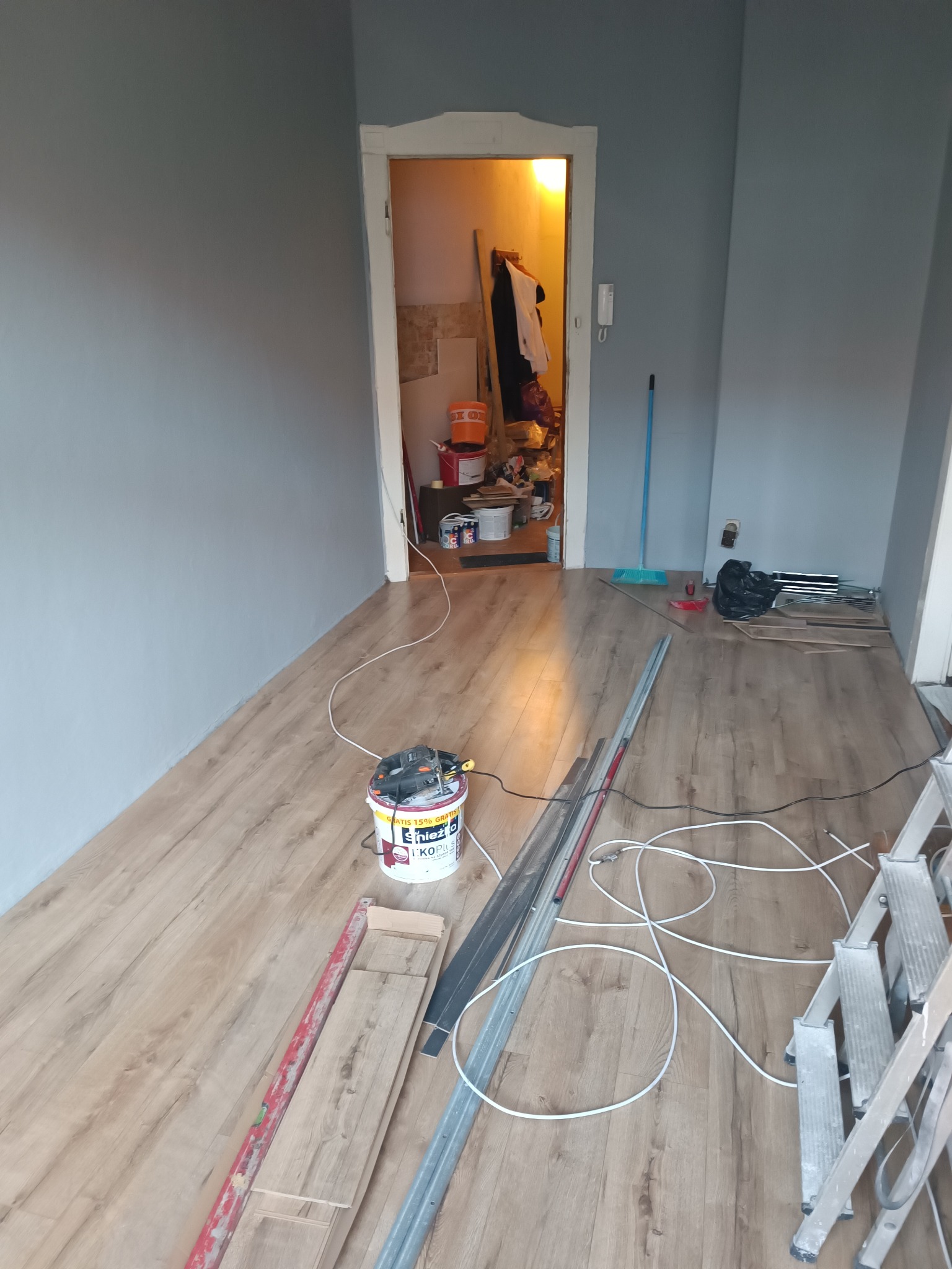 Pokój w trakcie remontu: świeżo pomalowane szare ściany, drewnopodobna podłoga, puszka farby, narzędzia, drabina i bałagan w przejściu do innego pomieszczenia.