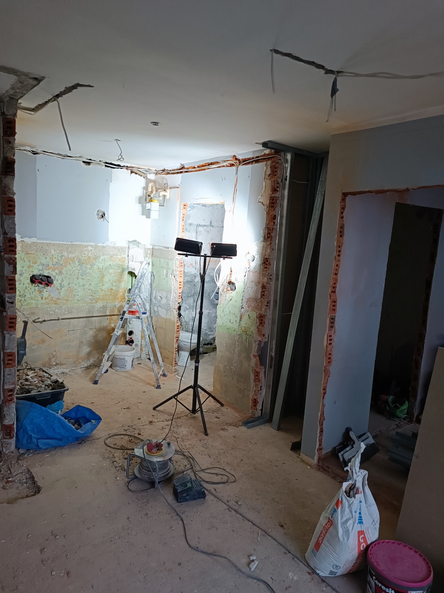 Wnętrze w trakcie remontu: odsłonięte cegły, przewody, drabina, worki z materiałami budowlanymi i reflektory na statywie oświetlające pomieszczenie.