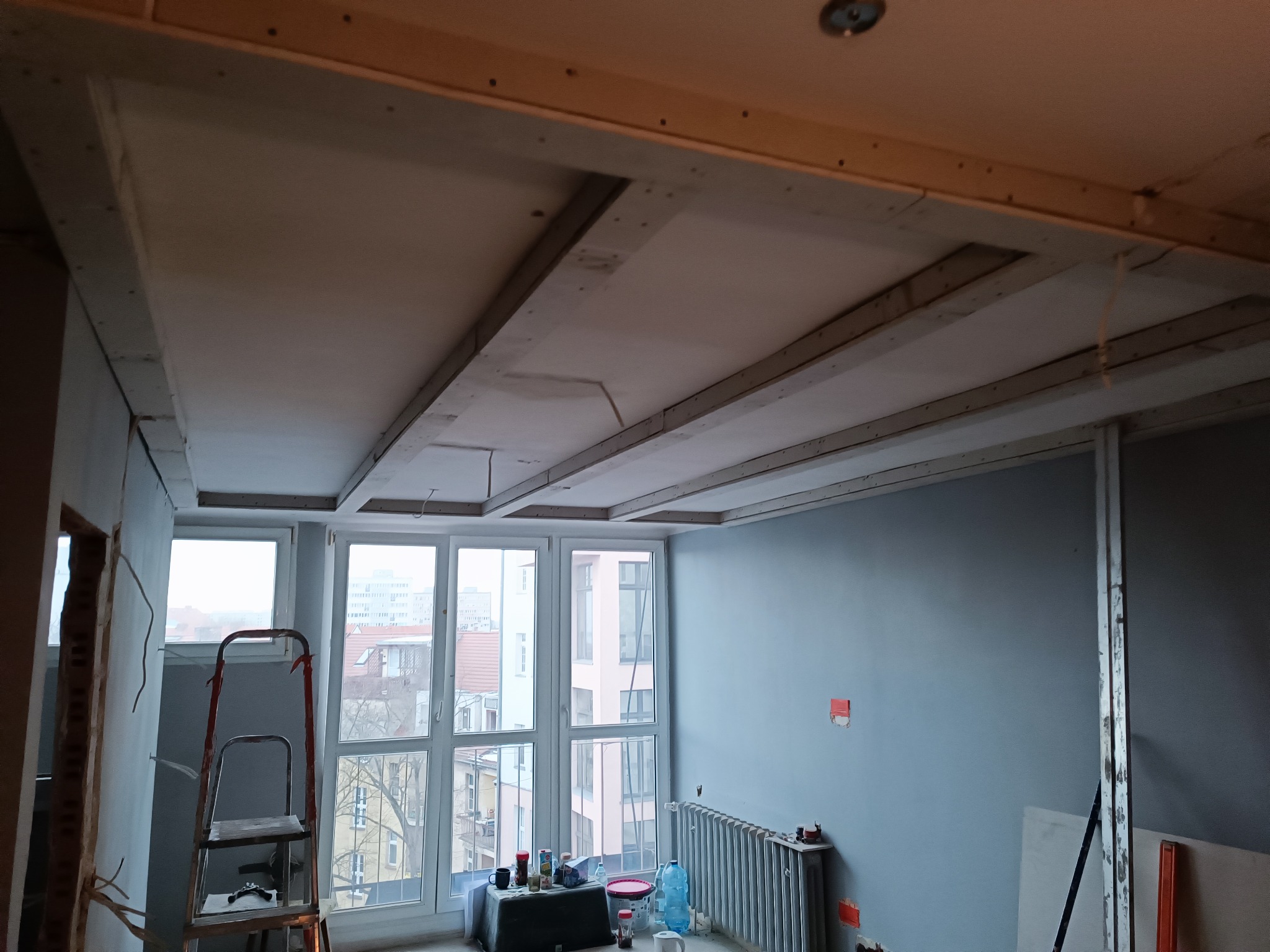 Sufit w trakcie montażu z widoczną konstrukcją drewnianą i częściowo zamontowaną płytą karton-gips, widok na okno i fragment pokoju z grzejnikiem.