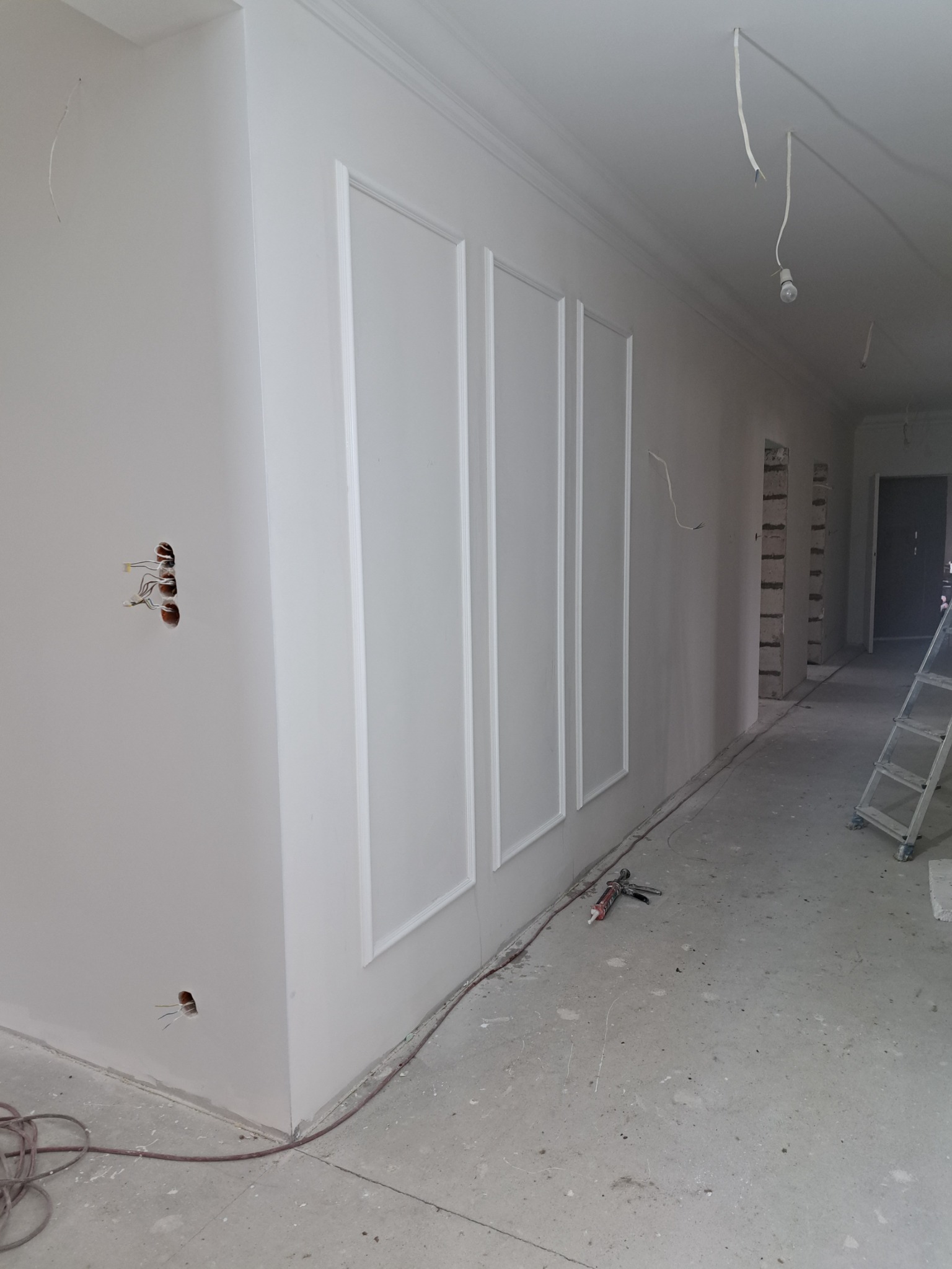 Korytarz w trakcie wykańczania: białe ściany z dekoracyjnymi listwami, widoczne przewody elektryczne, surowe podłoże i drabina w tle.