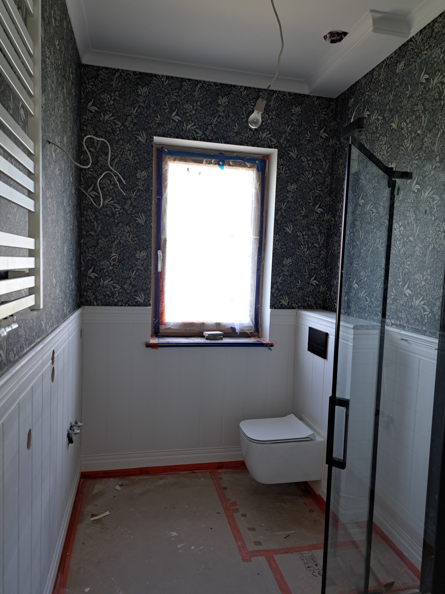 Wnętrze łazienki w trakcie prac wykończeniowych, widoczne: fragment kabiny prysznicowej z czarnymi profilami, podwieszana toaleta, grzejnik drabinkowy, okno zabezpieczone folią, wzorzysta tapeta...