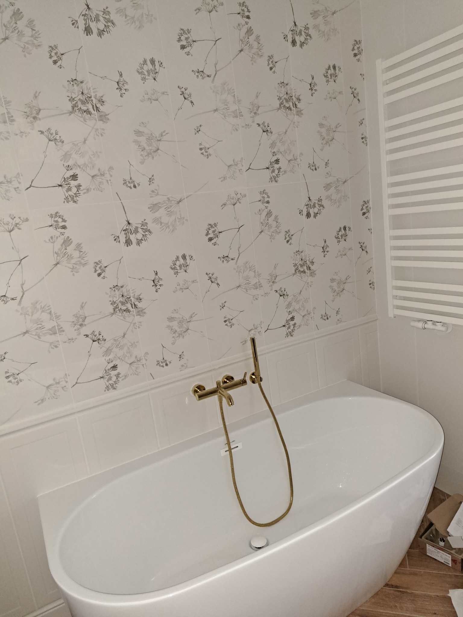 Biała wanna wolnostojąca z złotą armaturą na tle ściany wyłożonej płytkami z motywem kwiatowym i białym grzejnikiem drabinkowym