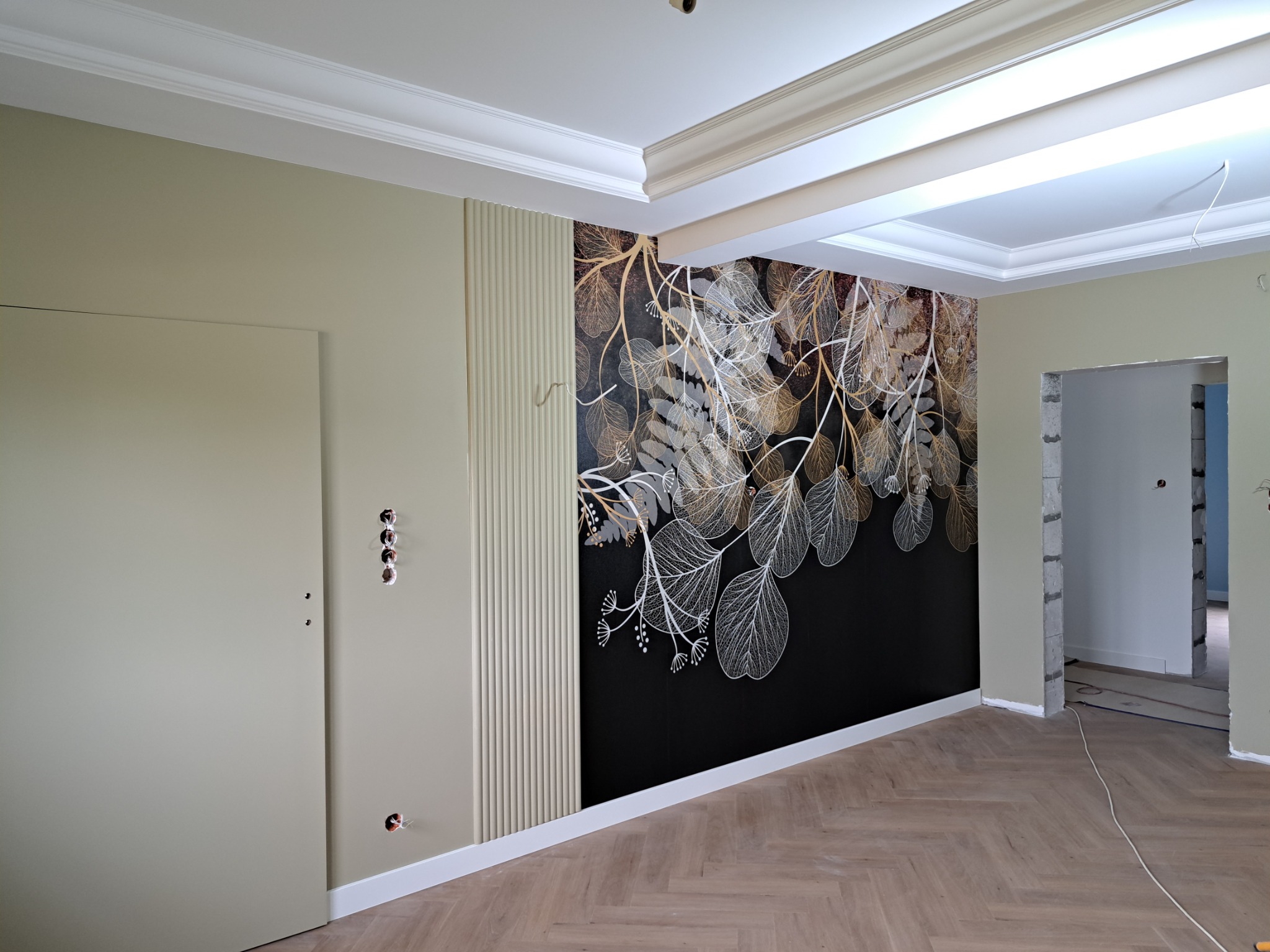 Wnętrze w trakcie wykańczania: ściana z dekoracyjnym panelem i tapetą w motywy roślinne, widoczne gniazdka elektryczne bez osprzętu, parkiet ułożony w jodełkę, sufit z ozdobną sztukaterią.