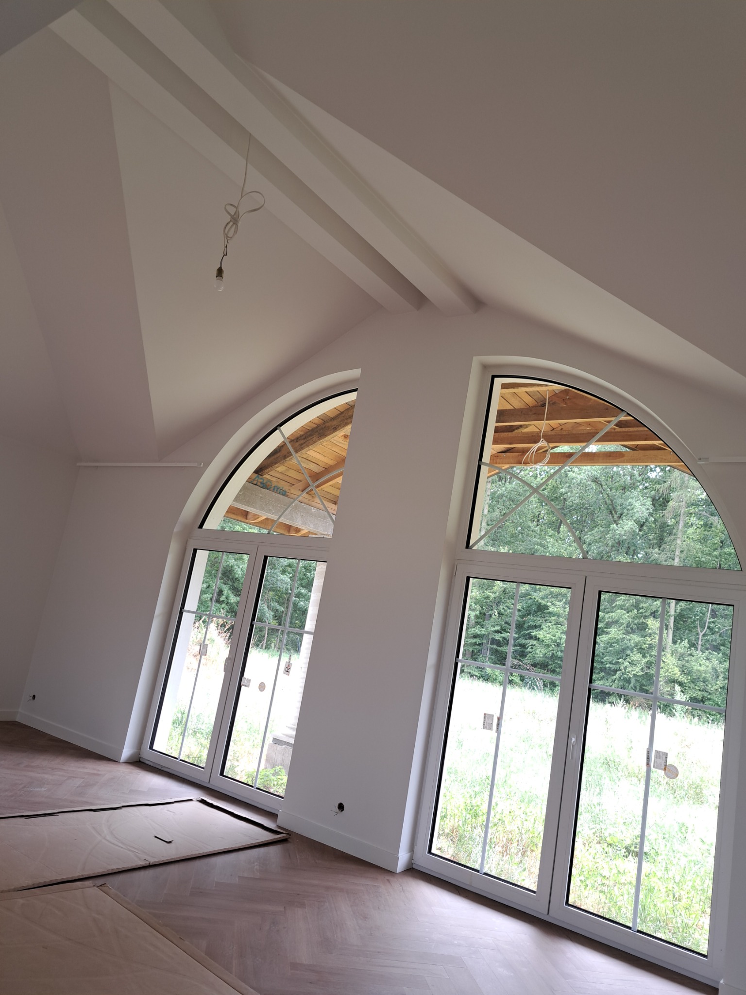 Poddasze z dwiema białymi, łukowymi oknami z widokiem na zieleń, drewniana podłoga w jodełkę zabezpieczona kartonem, surowe wykończenie ścian i sufitu, wisząca żarówka.