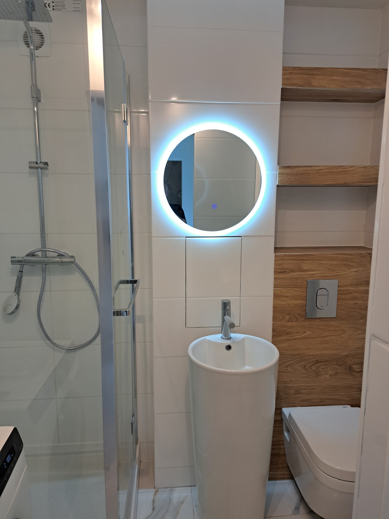 Nowoczesna łazienka z kabiną prysznicową, okrągłym lustrem z podświetleniem LED, umywalką na postumencie i drewnianymi półkami w zabudowie.