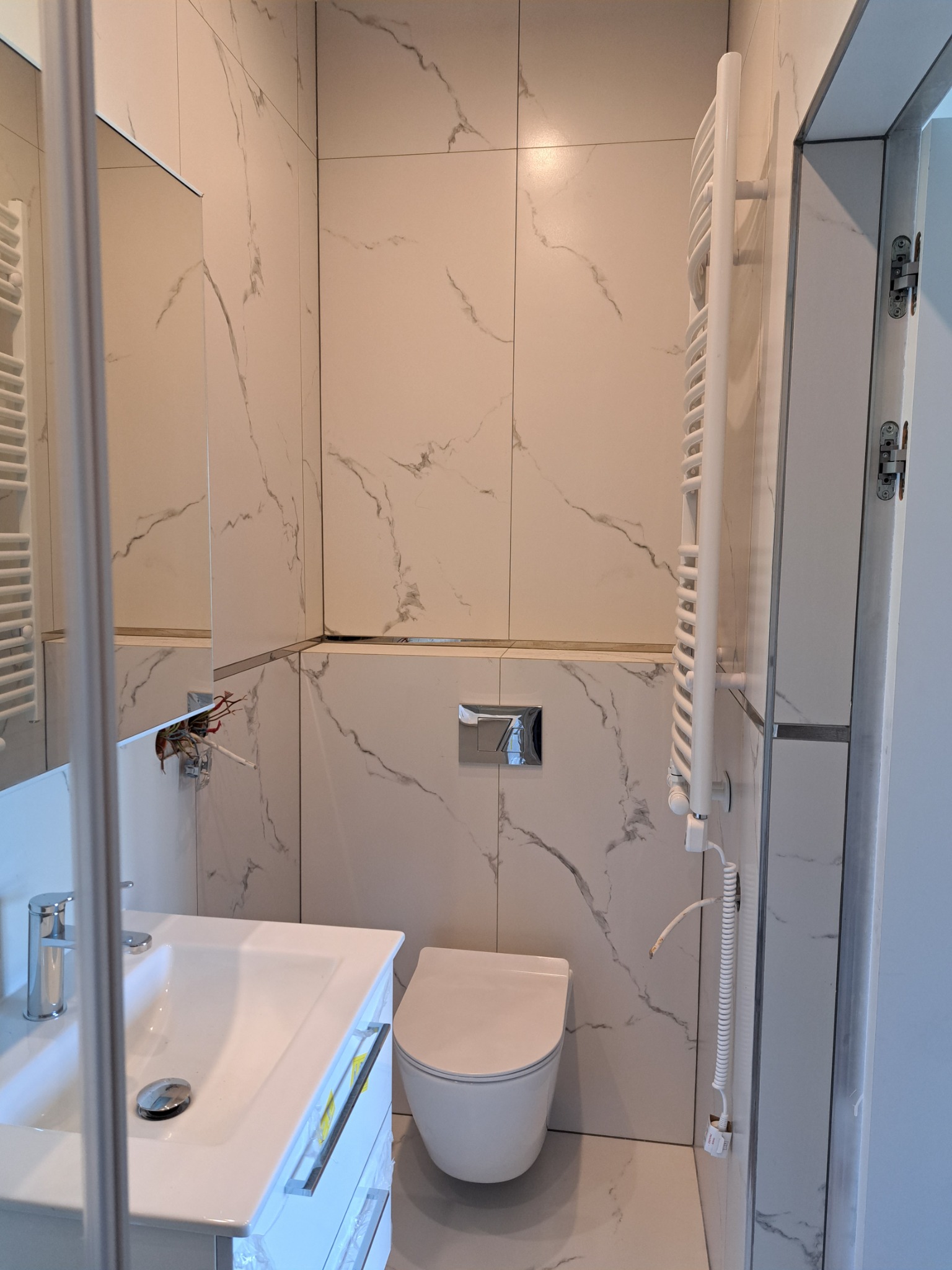 Nowoczesna łazienka z białymi płytkami imitującymi marmur, wisząca toaleta, umywalka z szafką i grzejnik drabinkowy, widoczne przewody elektryczne do podłączenia lustra.