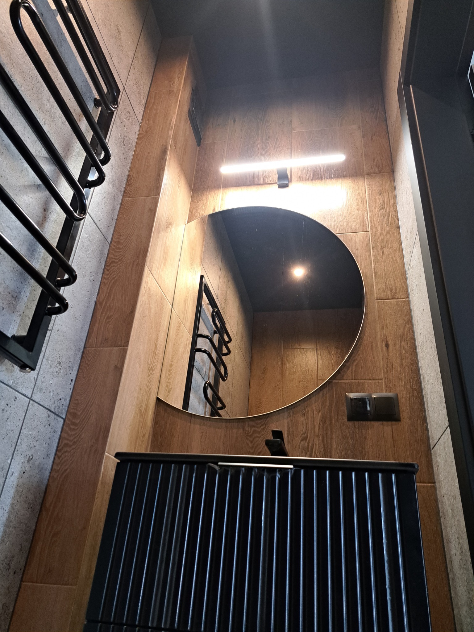 Nowoczesna łazienka z okrągłym lustrem podświetlanym listwą LED, czarną szafką umywalkową z pionowymi żłobieniami i grzejnikiem drabinkowym w kolorze czarnym, wykończona płytkami imitującymi drewno...