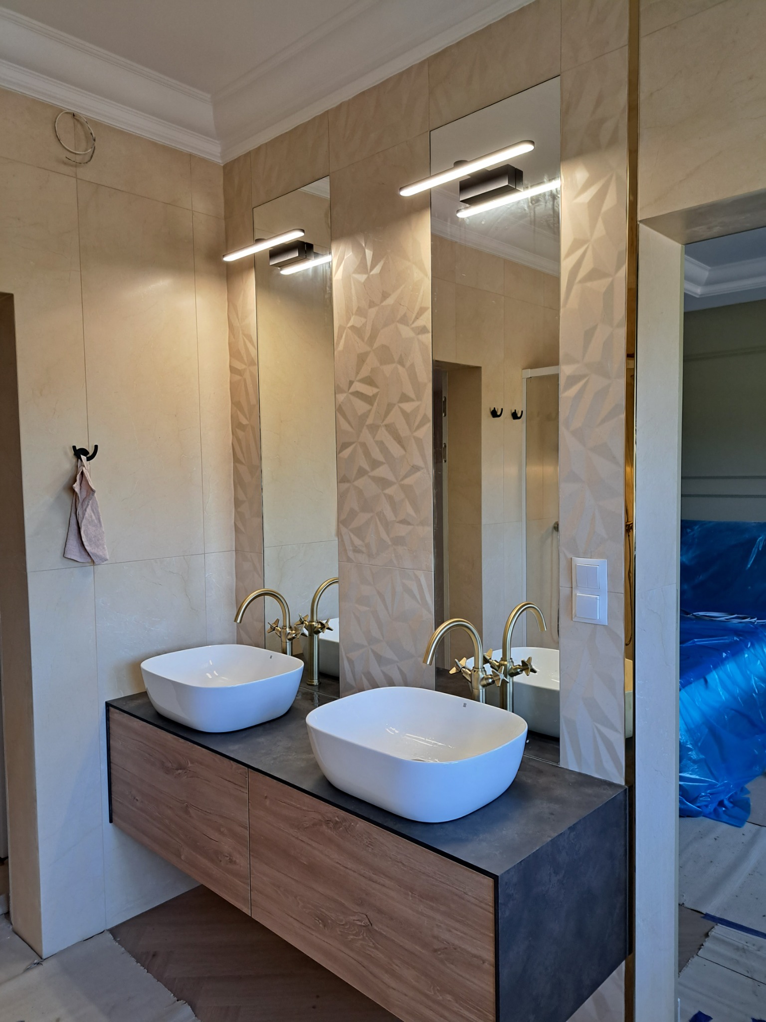 Nowoczesna łazienka z dwiema białymi umywalkami nablatowymi, złotą armaturą i podświetlanymi lustrami na tle beżowych płytek z geometrycznym wzorem. W tle widoczne prace wykończeniowe.