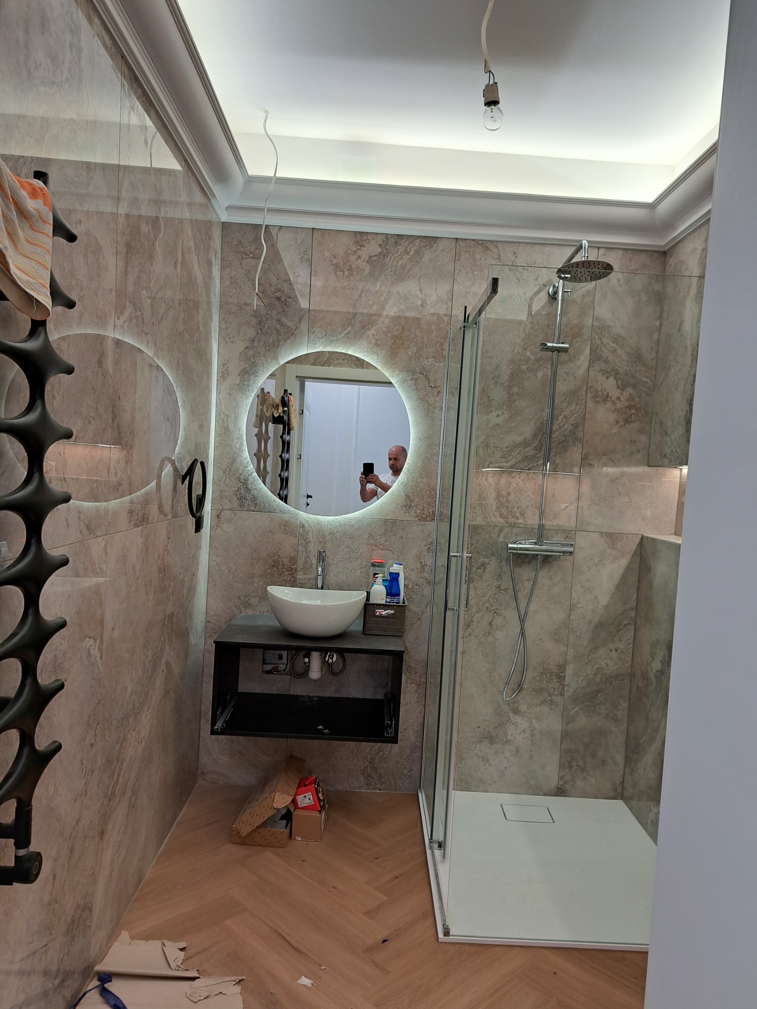 Ukończona łazienka z kabiną prysznicową, okrągłym lustrem podświetlanym LED, umywalką na czarnej szafce, grzejnikiem drabinkowym i płytkami imitującymi marmur. Na podłodze parkiet ułożony w jodełkę...