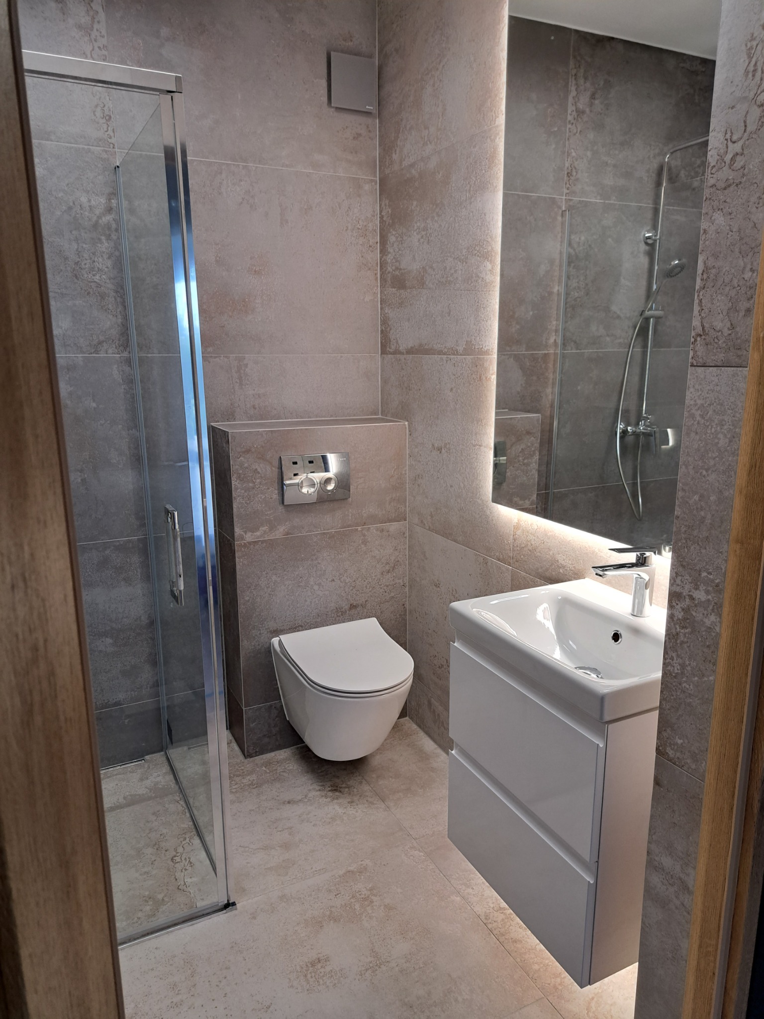 Nowoczesna łazienka z kabiną prysznicową, podwieszaną toaletą i umywalką z szafką, oświetlona lustrem LED i płytkami imitującymi kamień.