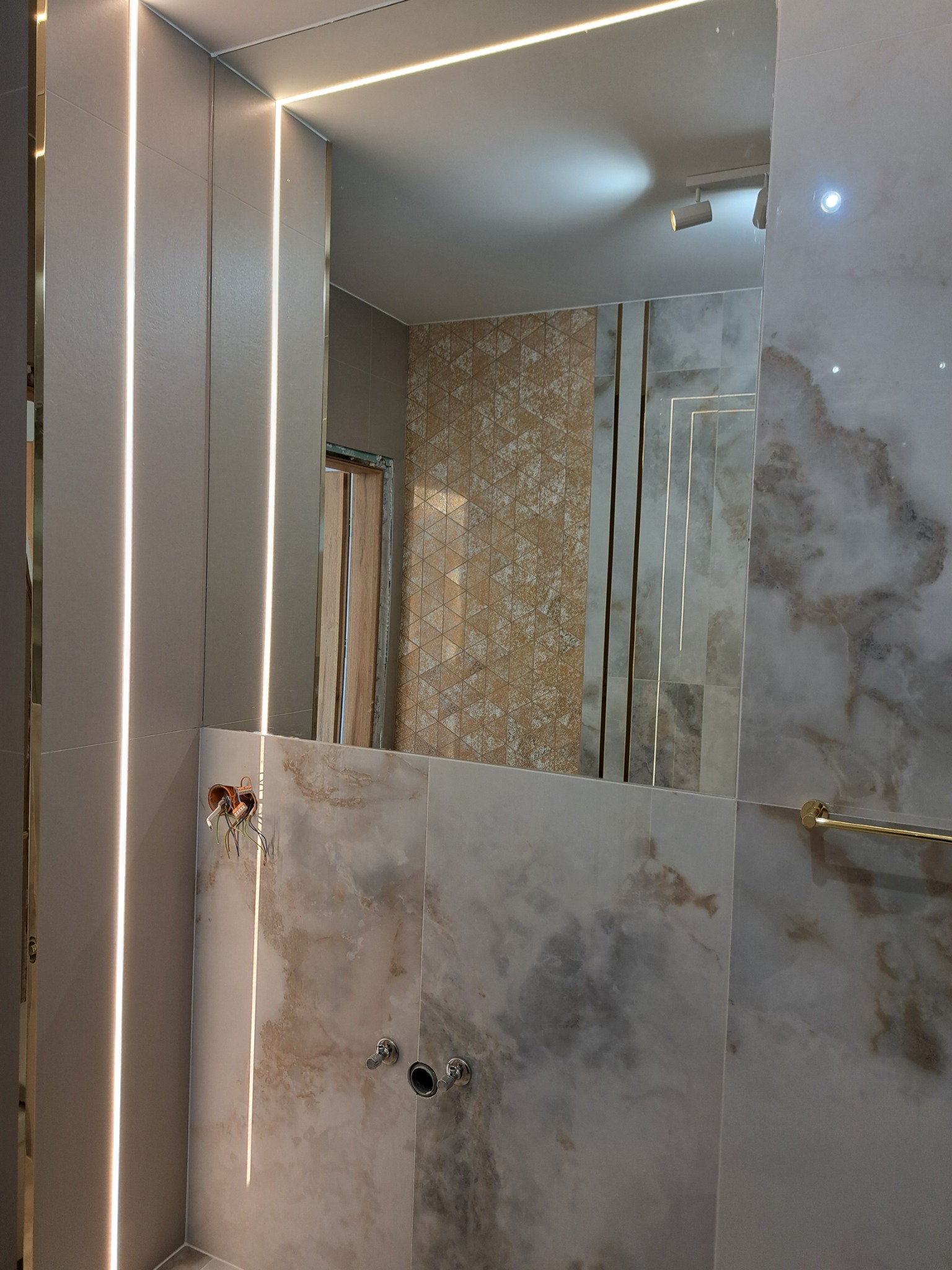 Nowoczesna łazienka w trakcie wykańczania, widoczne płytki imitujące marmur, oświetlenie LED w pionowych listwach, lustro z odbiciem fragmentu innej ściany z mozaiką, niezabezpieczone przewody...