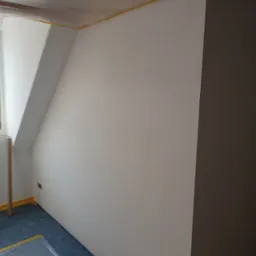 Świeżo pomalowane poddasze ze skosem, zabezpieczone folią malarską na podłodze i taśmą na suficie, jedna ściana w kolorze szarym, druga biała.