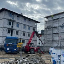 Budowa budynku wielorodzinnego w trakcie ocieplania, widoczne rusztowania, materiały izolacyjne i ciężarówka z dźwigiem teleskopowym na placu budowy.