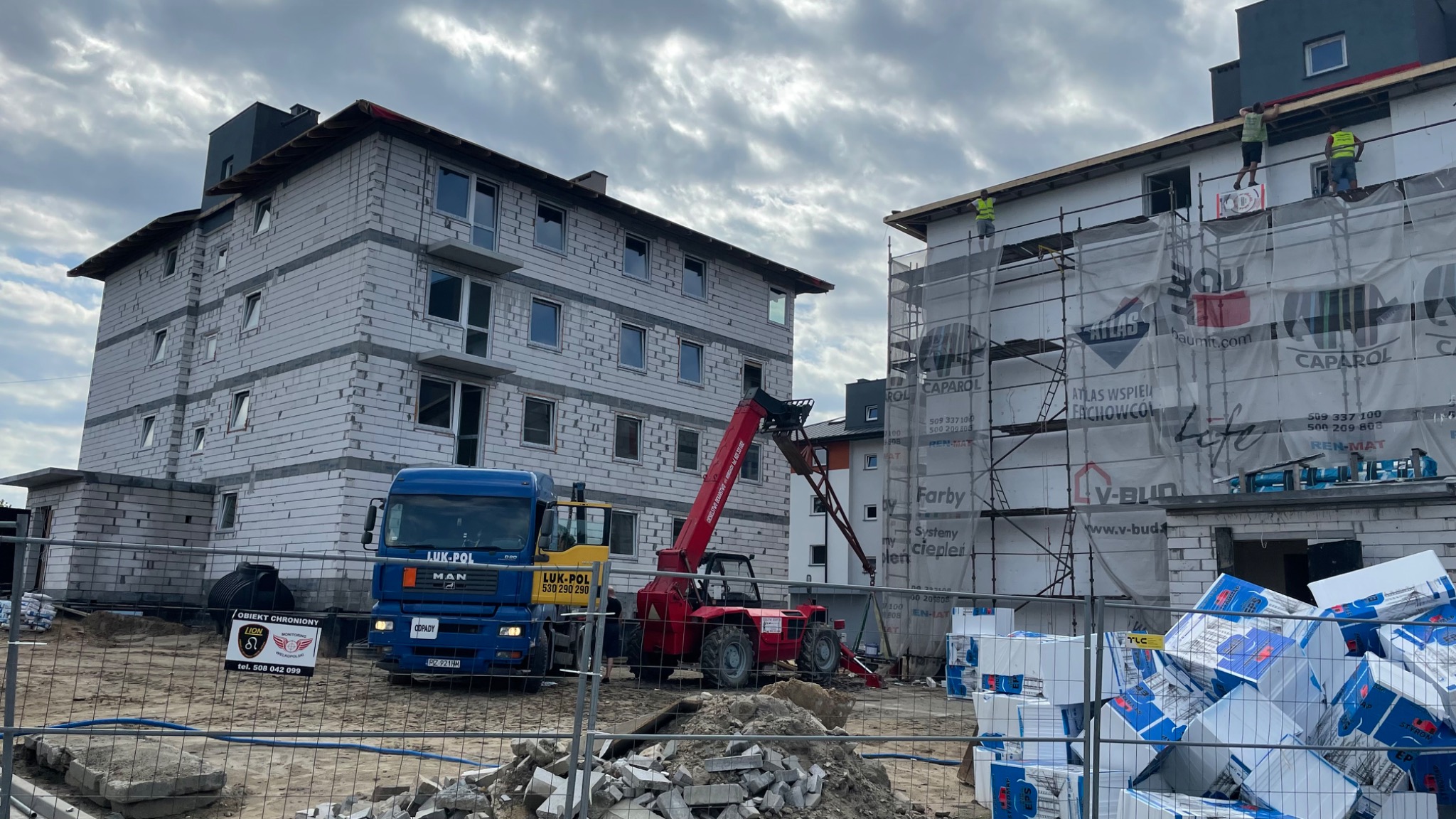 Budowa budynku wielorodzinnego w trakcie ocieplania, widoczne rusztowania, materiały izolacyjne i ciężarówka z dźwigiem teleskopowym na placu budowy.