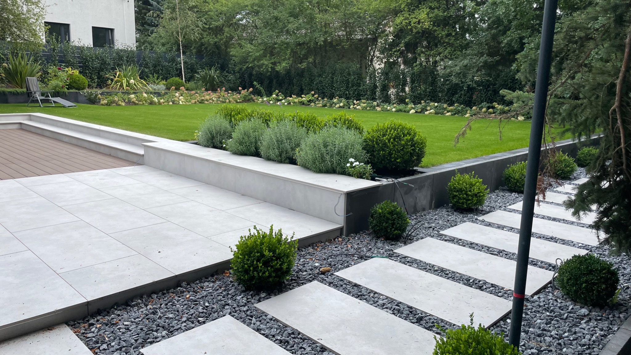 Nowoczesny ogród z tarasem z płytek, ścieżką z kamieni i równo przystrzyżonym trawnikiem, widoczne przewody elektryczne przy jednej z rabat.