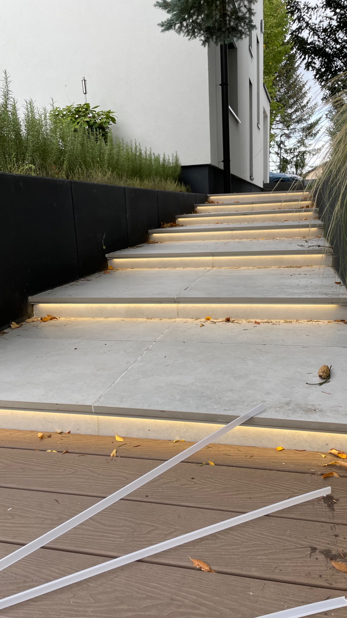 Zewnętrzne schody z podświetleniem LED wbudowanym w stopnie, prowadzące do nowoczesnego domu z roślinnością w tle, z widocznymi elementami montażowymi na drewnianym podeście.