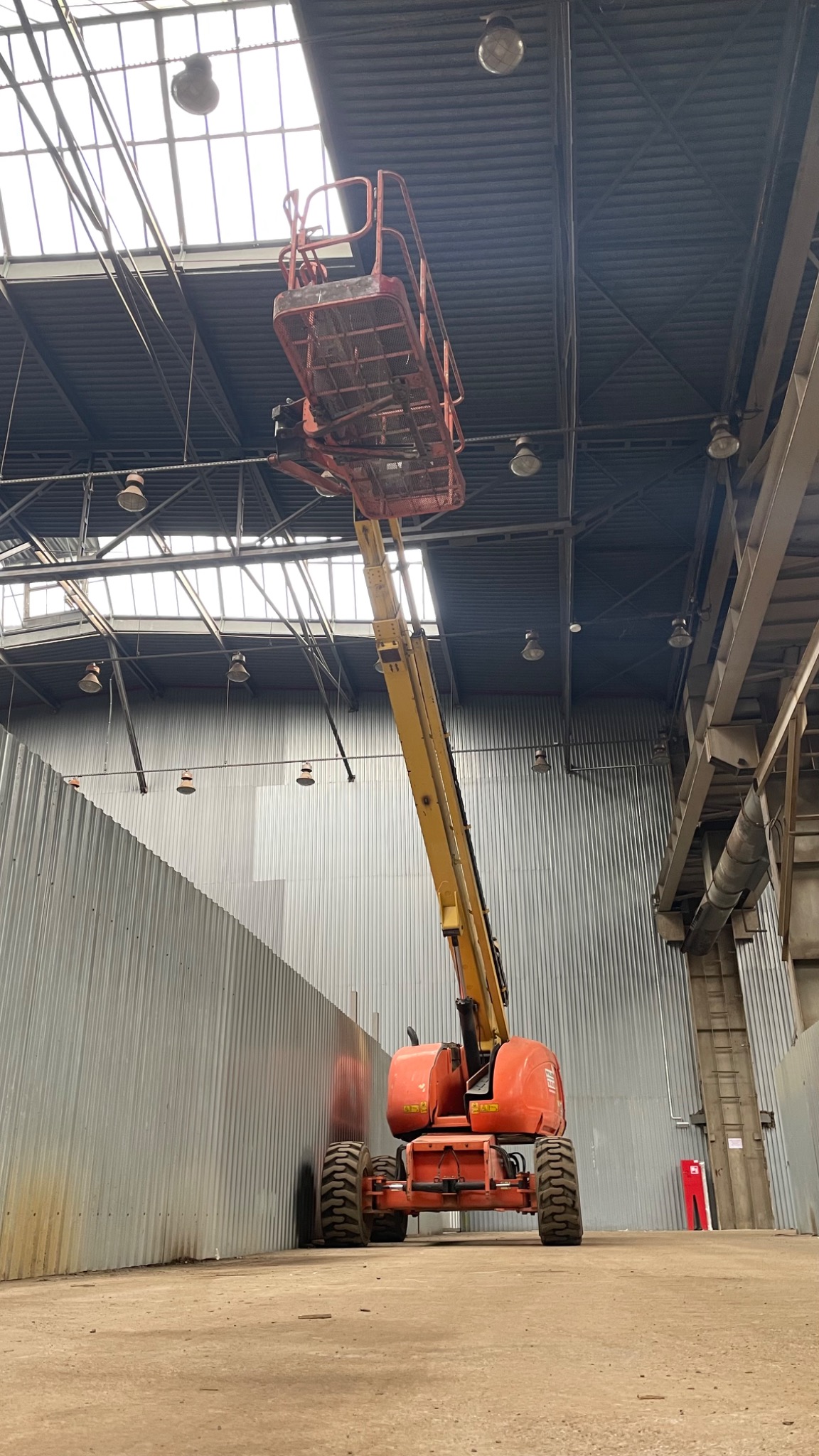 Pomarańczowo-żółty podnośnik koszowy w hali z blachy falistej, przygotowany do prac na wysokości, widoczne lampy pod sufitem.