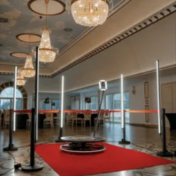 Oświetlona platforma obrotowa na czerwonym dywanie w eleganckiej sali balowej z żyrandolami i malowanym sufitem, przygotowana do sesji zdjęciowej.