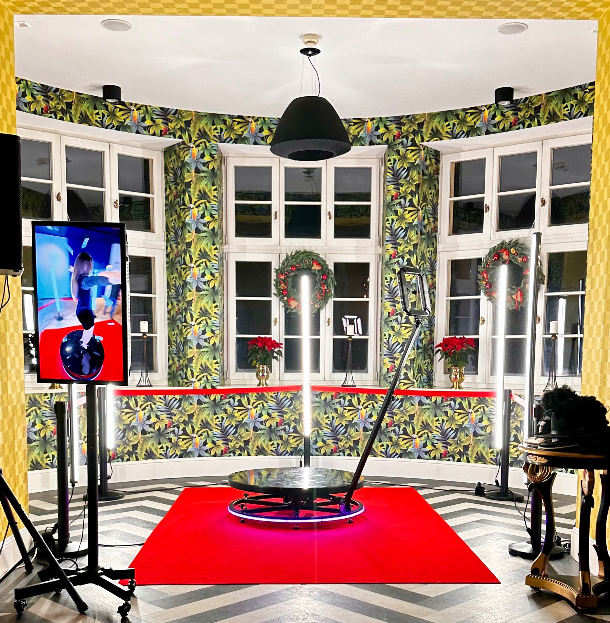 Wnętrze z zainstalowaną platformą obrotową 360 stopni na czerwonym dywanie, oświetlona lampami LED, w tle okna z wieńcami świątecznymi i tapeta w tropikalne liście.