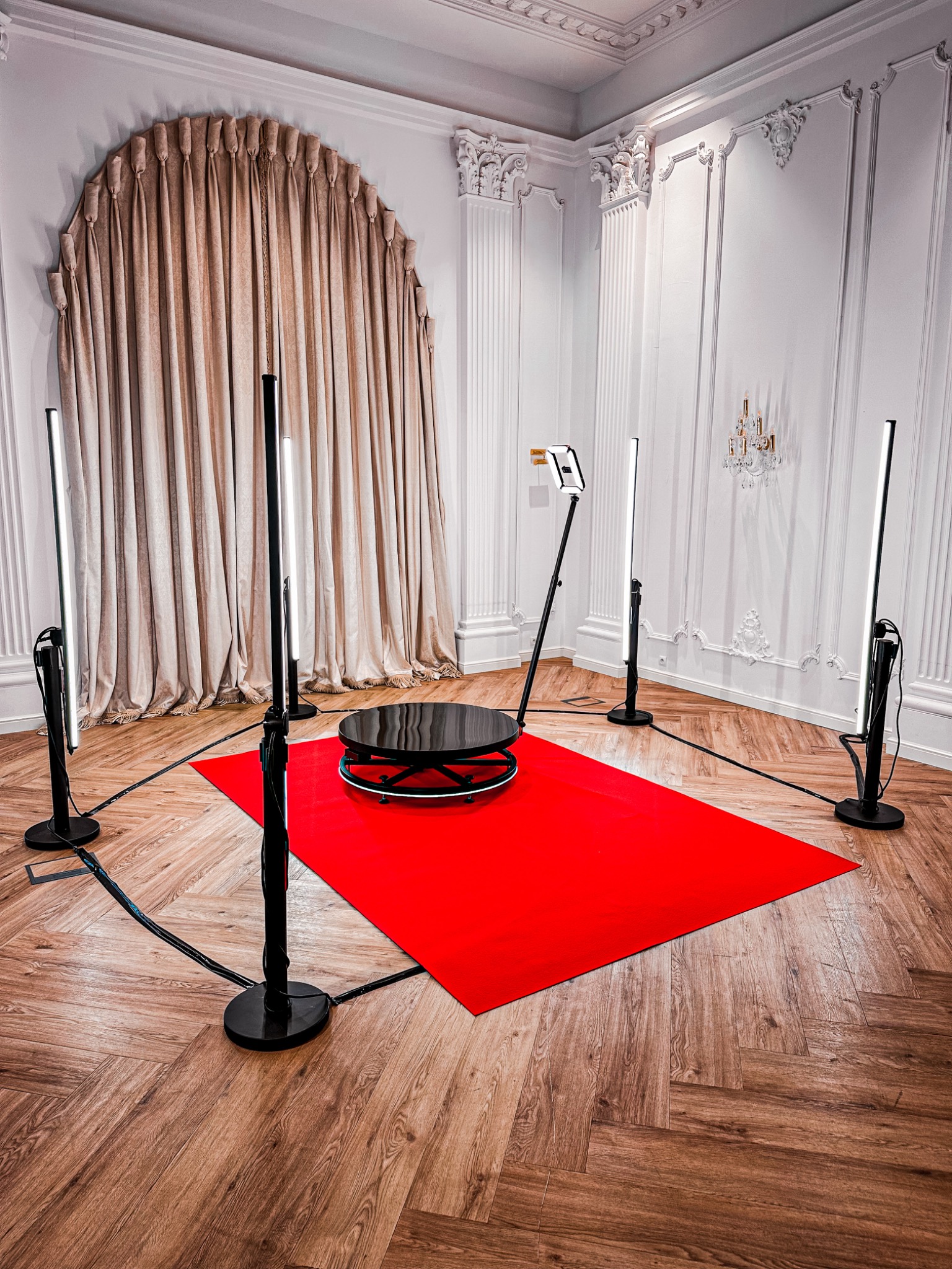 Obracana platforma do zdjęć 360 stopni z oświetleniem LED, ustawiona na czerwonym dywanie w eleganckim wnętrzu z dekoracyjnymi sztukateriami i żyrandolem.