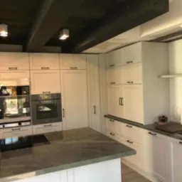 Elegancka kuchnia z białą zabudową i wyspą z blatem w odcieniach szarości. Czarne belki stropowe i minimalistyczne oświetlenie punktowe.