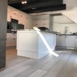 OMEGA TOMASZ KRĘCIOCH - Jasna kuchnia z wyspą, białe szafki i drewniane belki stropowe. Promień słońca oświetla podłogę z paneli. Nowoczesny design z elementami rustykalnymi.