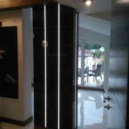 OMEGA TOMASZ KRĘCIOCH - Nowoczesna, czarna szafa wnękowa z oświetleniem LED, umieszczona w przestronnym holu z marmurową podłogą i widokiem na balkon. Minimalistyczny design, eleganckie wykończenie.