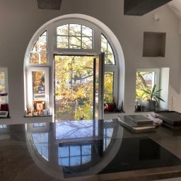 OMEGA TOMASZ KRĘCIOCH - Jasna kuchnia z oknem łukowym i widokiem na jesienne drzewa. Na blacie płyta indukcyjna i akcesoria. W tle przygotowane otwory na szafki.