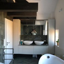 OMEGA TOMASZ KRĘCIOCH - Nowoczesna łazienka z marmurowymi ścianami, białymi szafkami pod umywalką i dużym lustrem. Widoczne drewniane belki stropowe i osoba robiąca zdjęcie w odbiciu lustra.