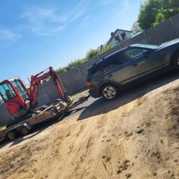 Nona trans - Ciemnoszary samochód osobowy z podpiętą przyczepą, na której znajduje się czerwona mini koparka. Usługa lawetowania w Ostrowie Wielkopolskim.