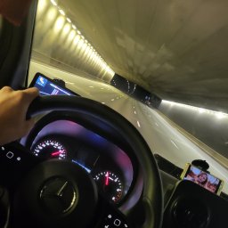 Nona trans - Wnętrze kabiny kierowcy podczas jazdy przez tunel, widok na deskę rozdzielczą, kierownicę z logo Mercedes, nawigację GPS i telefon z portretem kobiety, prędkościomierz wskazuje 100 km/h.