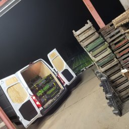 Nona trans - Biały van z otwartymi tylnymi drzwiami, załadowany skrzynkami z sadzonkami, stoi w magazynie obok sterty drewnianych palet. Widoczne oświetlenie magazynowe i częściowo pole uprawne w tle.