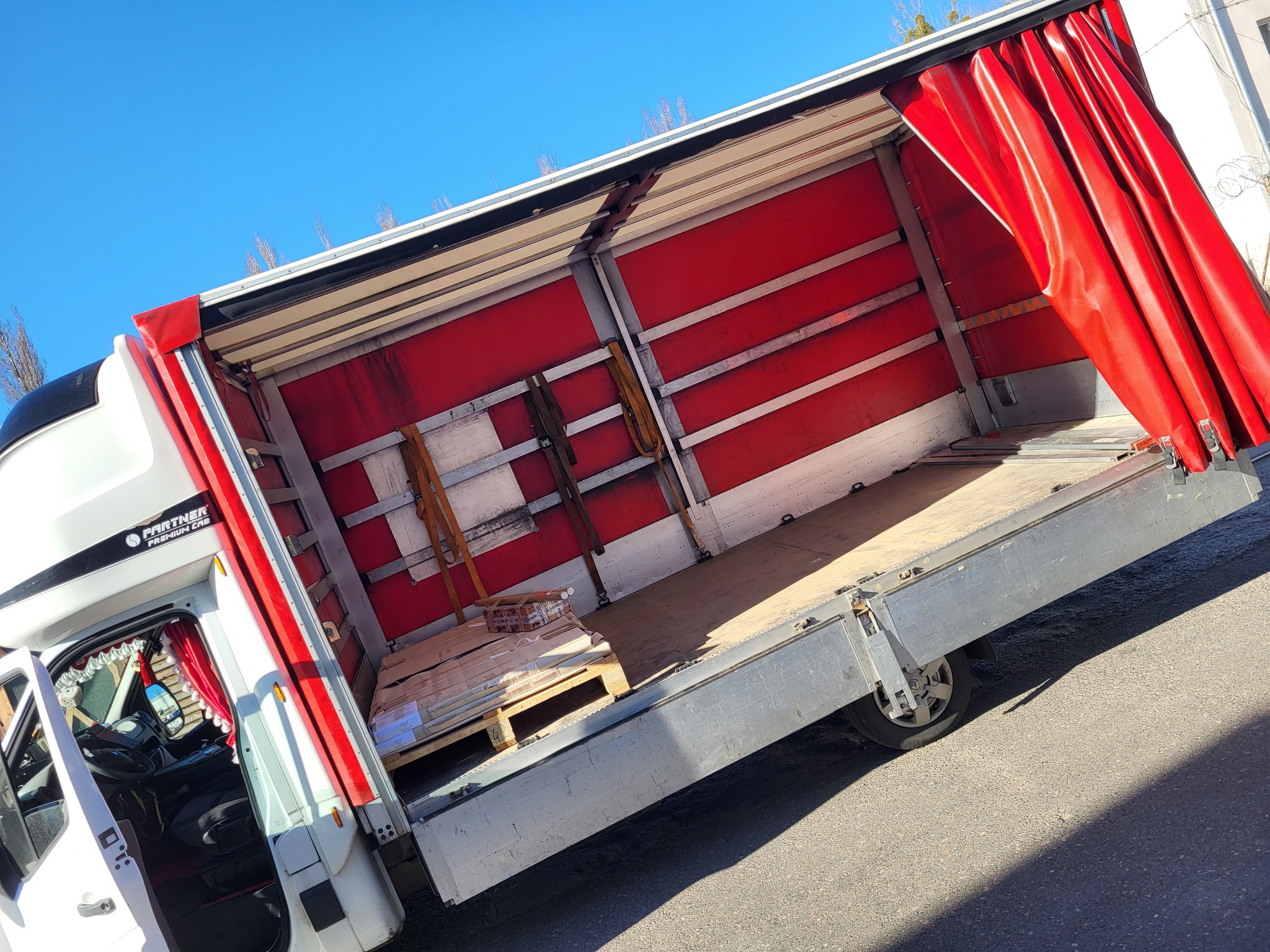 Otwarta przestrzeń ładunkowa busa z czerwonymi zasłonami, częściowo załadowana paleta z towarem, widok z boku na kabinę kierowcy.