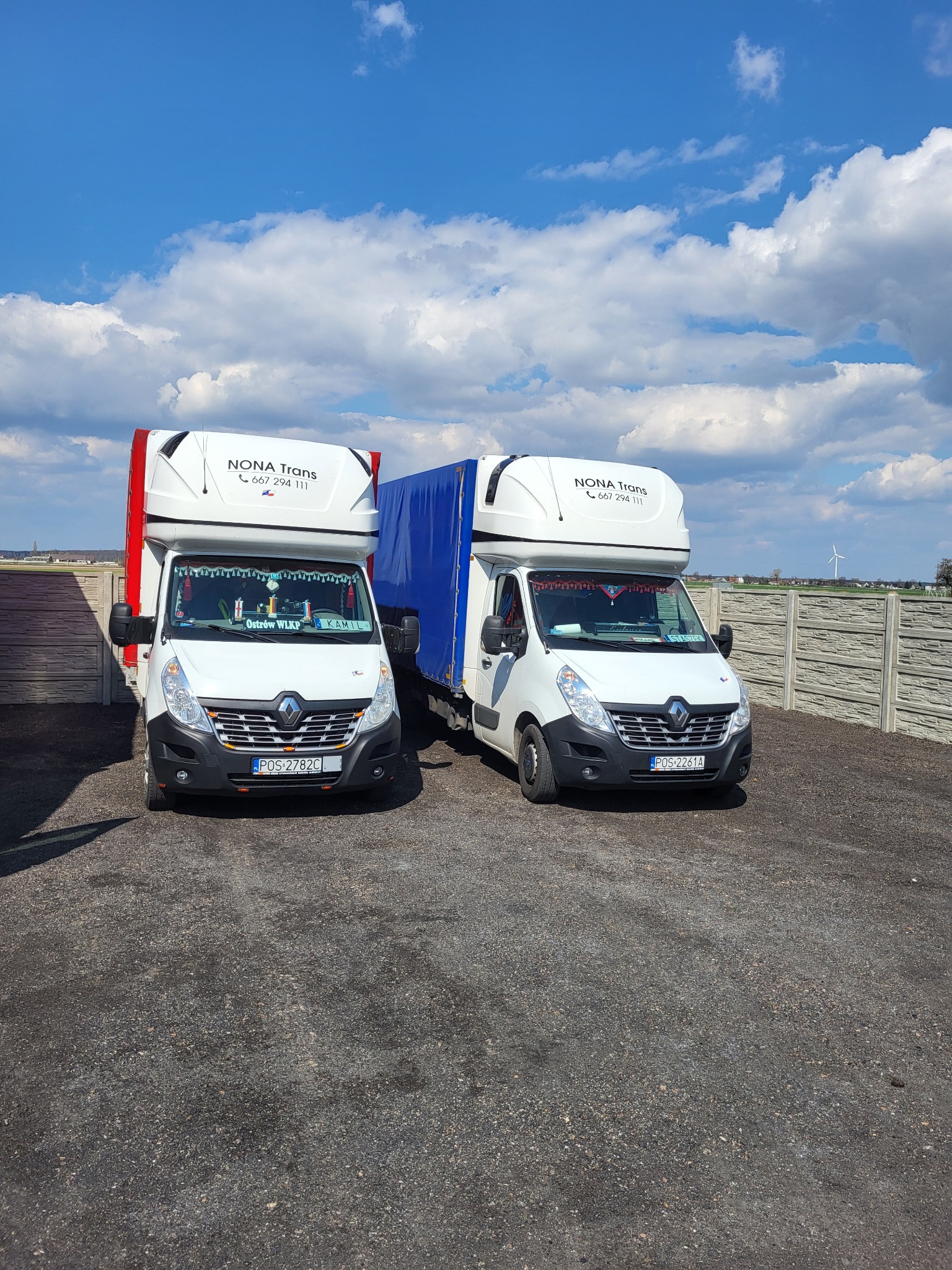 Dwa białe busy Renault Master z nadbudówkami, zaparkowane obok siebie na żwirowym placu, z widocznym logo firmy 'NONA Trans' i numerami telefonów na nadbudówkach, przed betonowym ogrodzeniem...