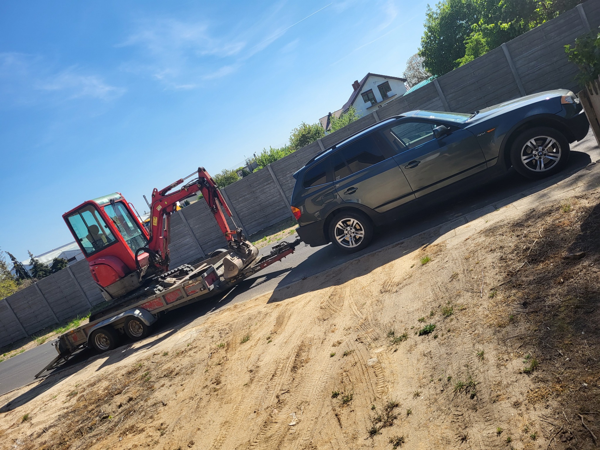 Ciemnoszary samochód osobowy z podpiętą przyczepą, na której znajduje się czerwona mini koparka. Usługa lawetowania w Ostrowie Wielkopolskim.