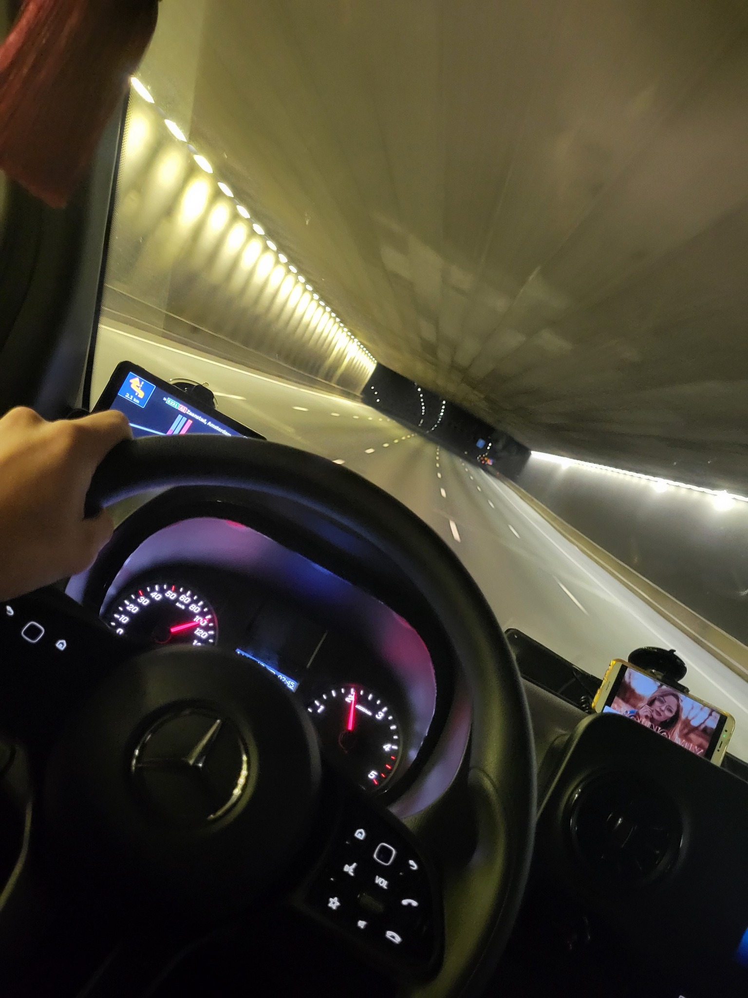 Wnętrze kabiny kierowcy podczas jazdy przez tunel, widok na deskę rozdzielczą, kierownicę z logo Mercedes, nawigację GPS i telefon z portretem kobiety, prędkościomierz wskazuje 100 km/h.