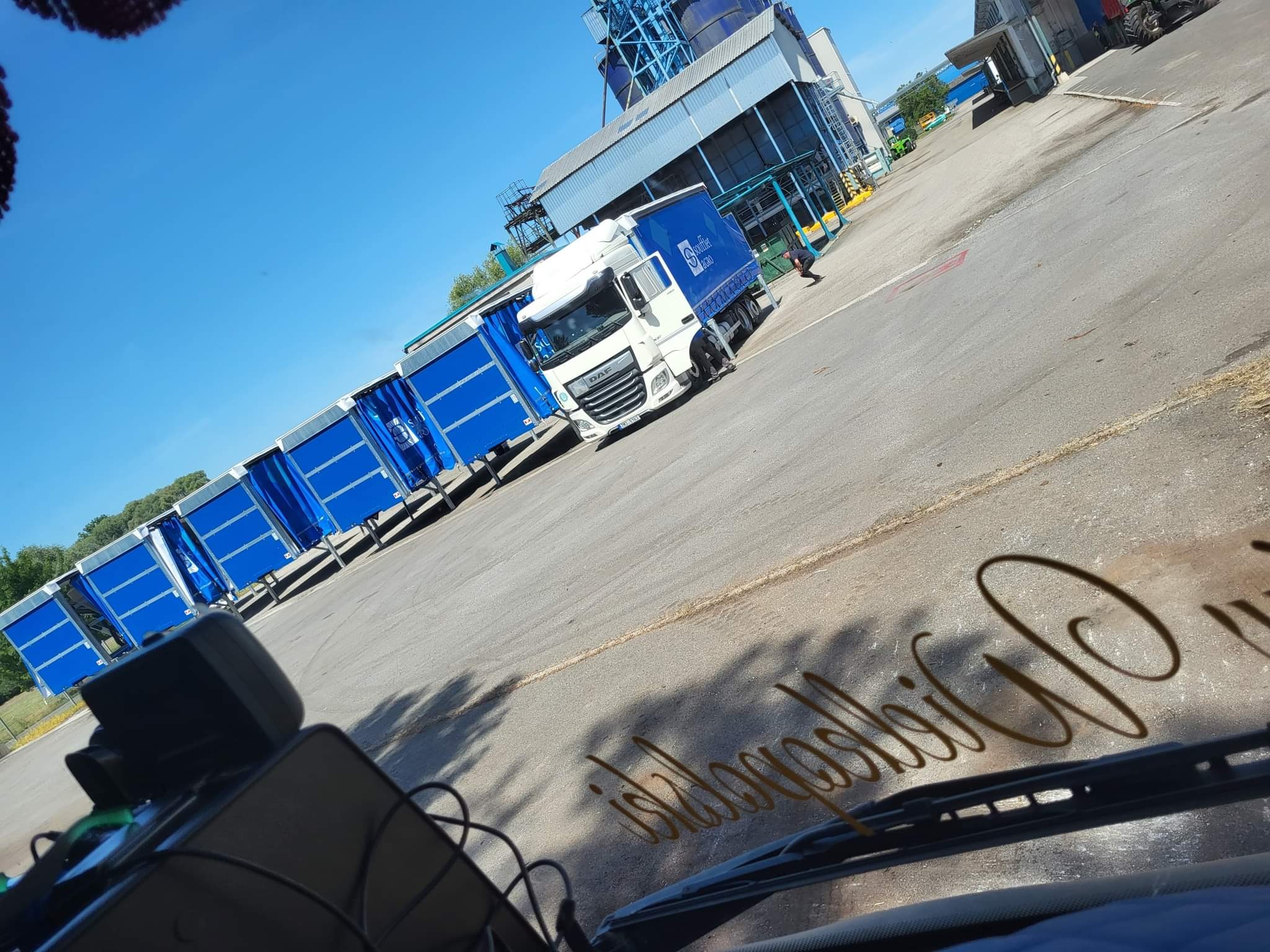 Ciężarówka z przyczepą z plandekami w kolorze niebieskim, zaparkowana na placu manewrowym z budynkami przemysłowymi w tle, widok z kabiny kierowcy z widocznym odbiciem napisu na przedniej szybie.