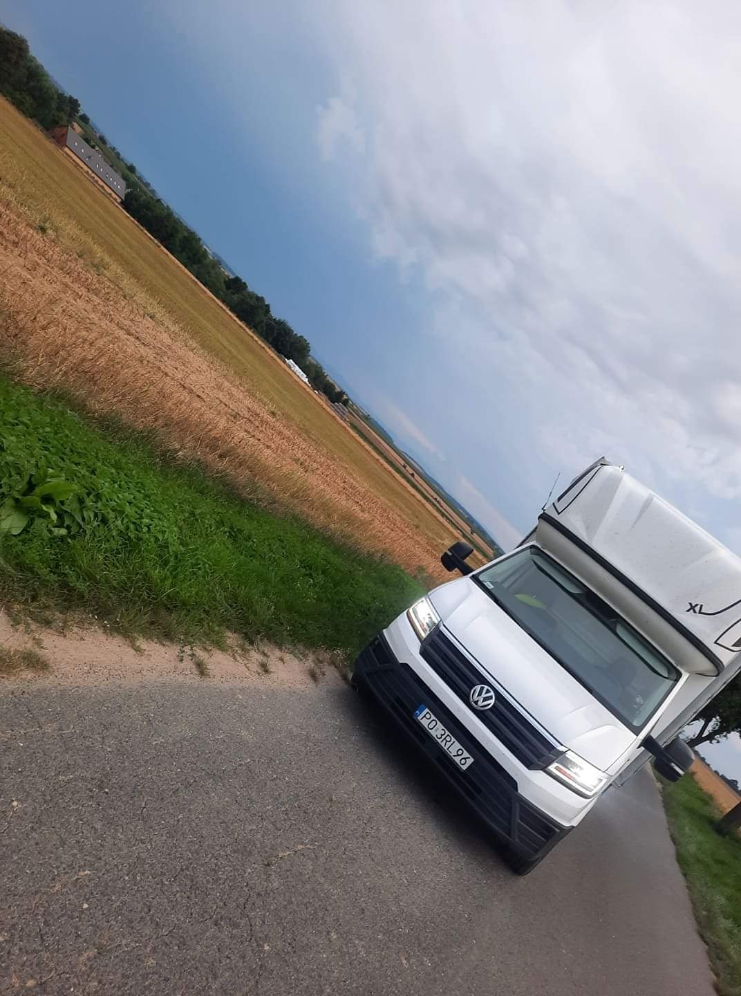 Biały samochód dostawczy Volkswagen z zabudową typu kontener na tle pola i pochmurnego nieba, widziany z niskiej perspektywy.