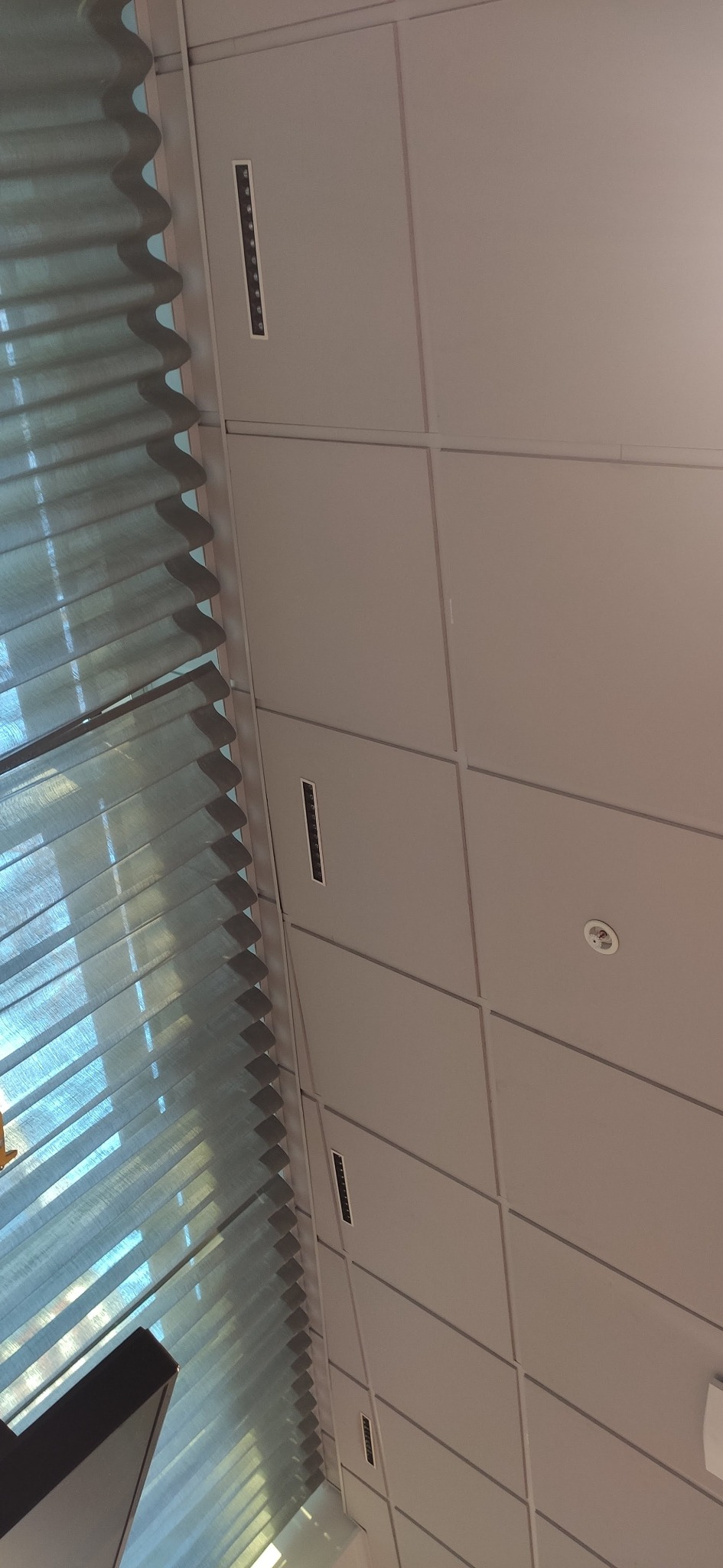 Kasetonowy sufit podwieszany w biurze z wbudowanymi kratkami wentylacyjnymi i oświetleniem, widok z dołu, z fragmentem żaluzji okiennych.